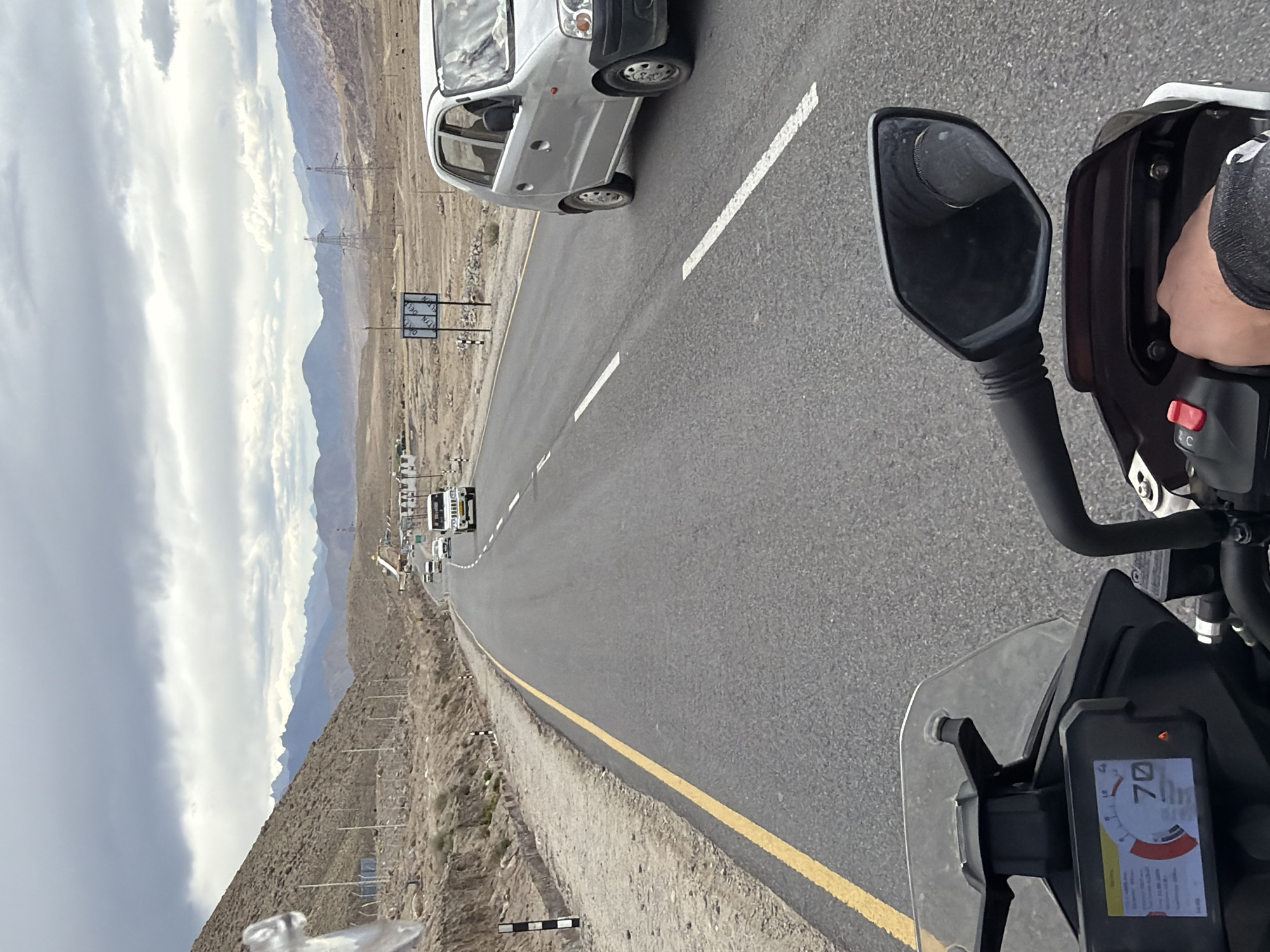 The image captures a first-person perspective of riding a motorcycle on a paved road in a mountainous, arid region. The road is asphalt with a yellow line marking the edge and white dashed lines indicating lanes. Several vehicles, including a white SUV and a silver hatchback, are visible on the road ahead. The landscape is characterized by barren, rocky hills and distant mountains under a cloudy sky. In the foreground, the motorcycle's dashboard displays a speed of 70, and a rider's hand is visible on the handlebars. The location is identified as Chumkar, India. The scene appears to be a daytime journey on a highway.