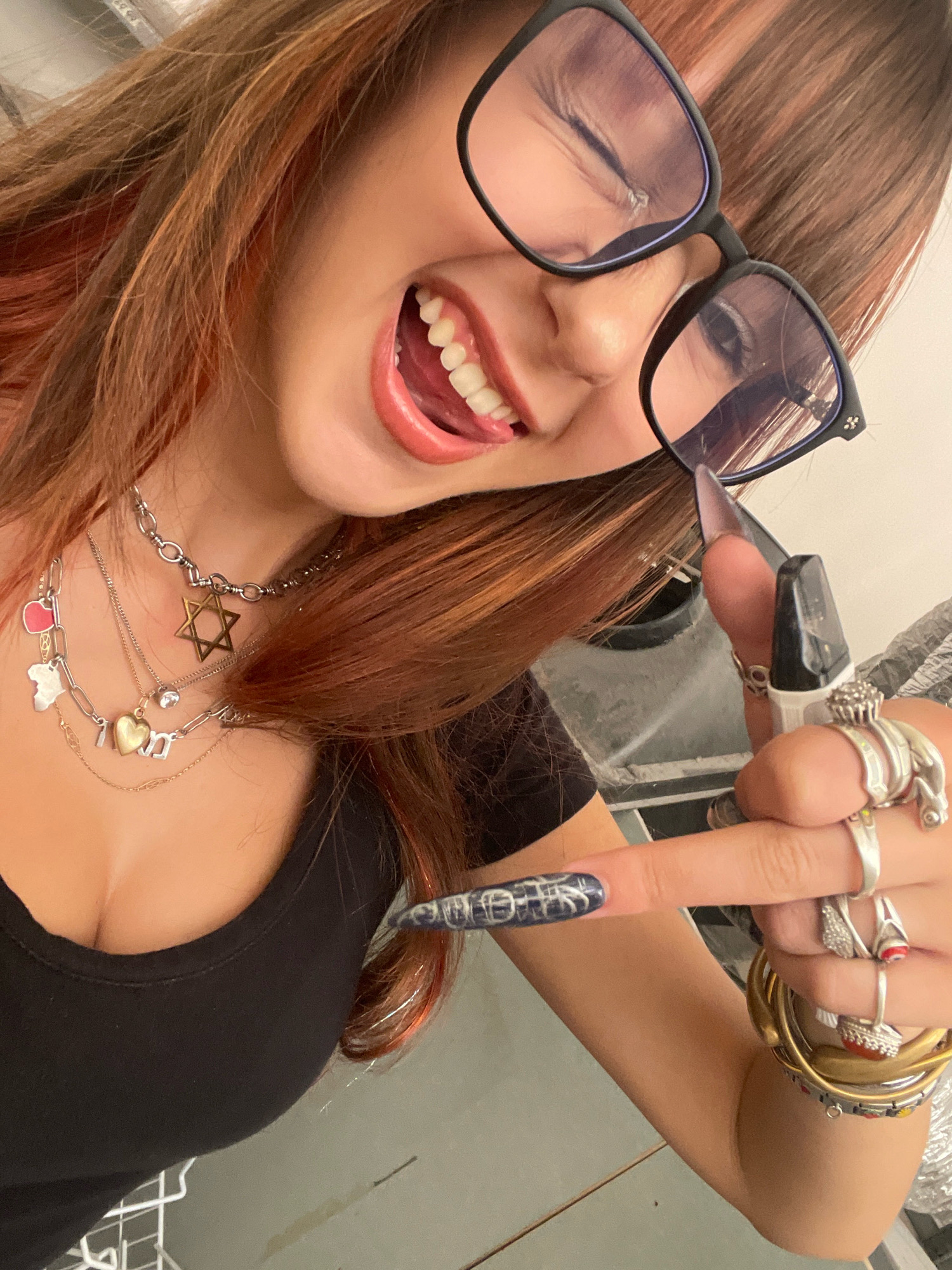 The image shows a close-up, high-angle shot of a young woman's upper body, taken indoors. She is wearing a black top and multiple necklaces, including one with a Star of David pendant and others with charms. She has long, reddish-brown hair and is wearing glasses. Her mouth is open, and her tongue is visible. She is holding a vape pen in her right hand, which is adorned with several rings and long, dark nails with a blue and white pattern. Her left hand is also visible, with rings and long nails. The background appears to be a domestic or industrial setting, with hints of metal and possibly shelving. There are no specific indicators of the location being in Raanana, Israel.