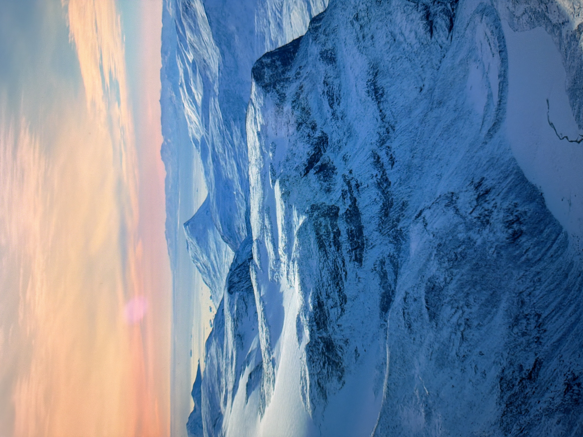 This is a landscape photograph of snow-covered mountains and a body of water under a pastel-colored sky. The sky transitions from pale orange and pink near the horizon to pale blue overhead. The mountains are rugged and appear to be glacial, with sharp peaks and deep valleys. Snow covers the tops and slopes of the mountains, creating a stark contrast with the darker rock formations visible in some areas. In the distance, a calm body of water, possibly a fjord or sea, is visible, with a few small ice floes or islands scattered across its surface. The overall impression is one of a cold, remote, and serene natural environment. There is no indication that this image is related to the city of Kyiv, Ukraine.