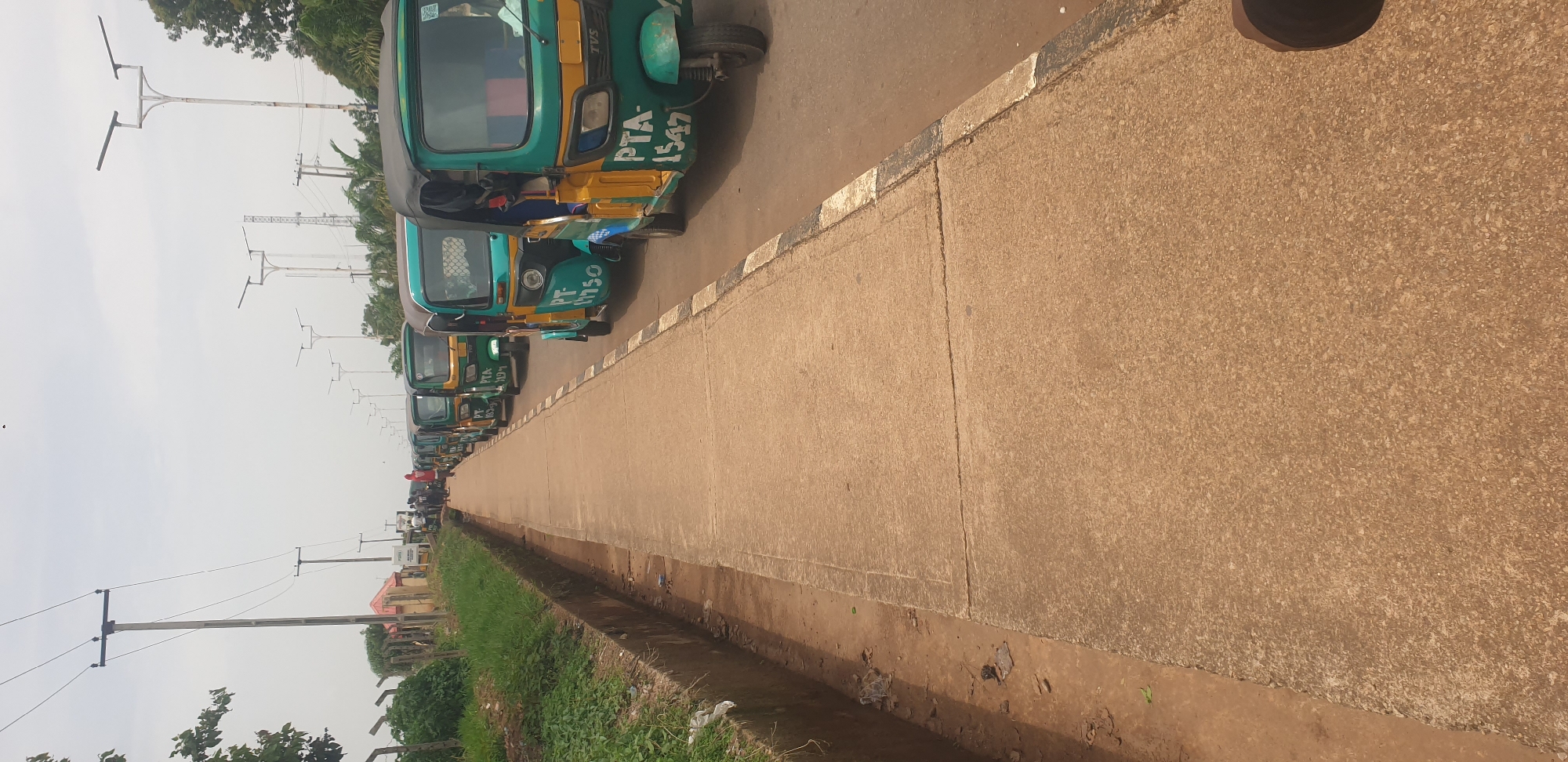 The image depicts a line of three-wheeled vehicles, commonly known as tricycles or "keke napeps," parked along a paved road. The vehicles are predominantly green and yellow and have white lettering on their sides, indicating registration numbers such as "PTA 1547" and "PT 1750." Several individuals are visible standing or walking near the vehicles, with one person's leg and foot visible in the lower right corner of the frame.
The scene is located in Barakin Lerawi, Nigeria. A concrete sidewalk runs parallel to the road, separated by a narrow drainage ditch filled with dirt and debris. Beyond the sidewalk, there is a grassy area and a fence. In the background, a utility pole with electrical wires is prominent, along with several modern streetlights and trees. The sky is overcast, suggesting it might be a cloudy day. The overall impression is of a typical street scene in a Nigerian city, with commercial tricycles waiting for passengers or parked.