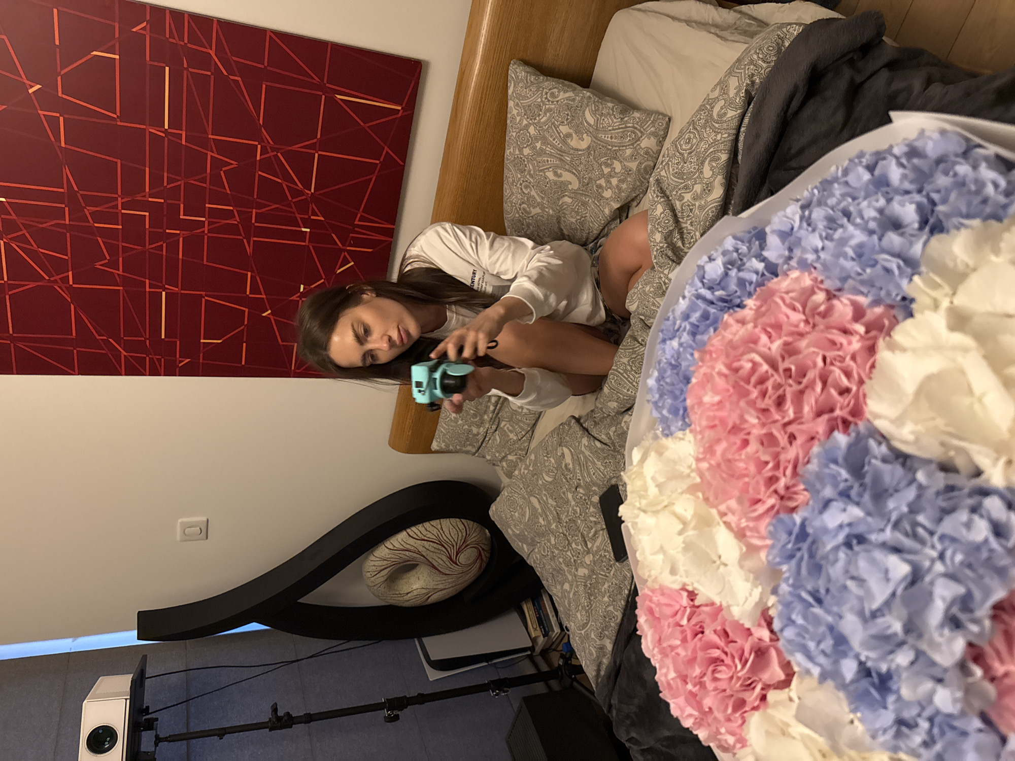 A young woman with long, dark hair is depicted lying on her side on a bed, looking down at a small, turquoise-colored camera she holds in her hands. She is wearing a white long-sleeved top that has some visible text, including the partial word "ENTURY." Her expression is focused and calm.
The setting appears to be a bedroom or private living space. The bed has a wooden headboard, light grey patterned pillows, and dark grey bedding. Resting prominently on the bed to the right is a large bouquet of flowers, arranged in sections of pink, blue, and white petals, creating a vibrant display.
Behind the woman, on the wall, hangs a large piece of abstract art featuring a dark red background with an intricate network of lighter red and orange geometric lines. Below the bed, to the left, a white projector is set up on a stand, alongside a black speaker. A white PlayStation 5 console is also visible near the base of the bed, beneath a decorative stand. This stand features a dark, curved design and holds a unique, oval-shaped white sculpture with a swirling red pattern. A light switch is visible on the wall further to the left. The indoor lighting suggests it is likely evening or night. The scene captures a quiet, personal moment.