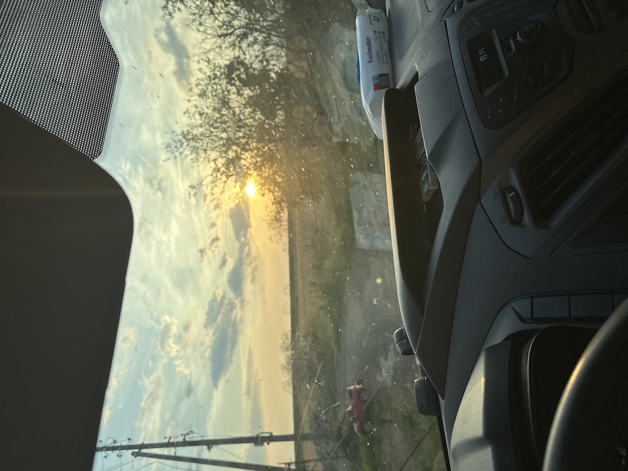 This image, taken from inside a vehicle in Mlekarevo, Bulgaria, captures a rural landscape at sunset. The view through the windshield shows a dusty road leading into a field under a sky filled with scattered clouds and the setting sun. On the left, a power line pole stands prominently. A small red object, possibly a toy car, is visible on the side of the road. The vehicle's dashboard is in the foreground, with a pack of cigarettes in a storage compartment and a pack of "Freshmaker" wet wipes on top. The sun's rays are casting a warm glow across the scene, creating lens flare and highlighting dust particles on the windshield. The overall impression is one of a quiet, rural setting during a beautiful sunset.