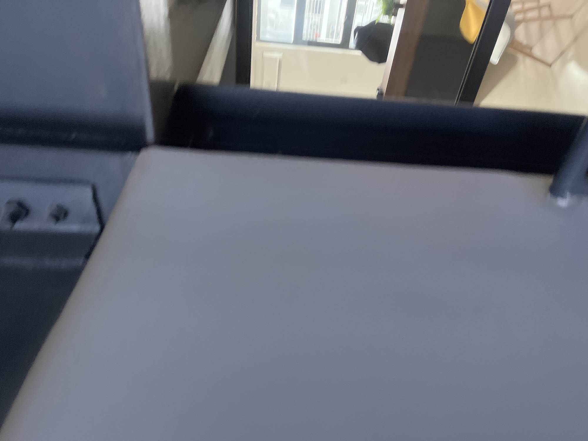 The image shows a close-up of a light gray surface in the foreground, possibly a wall or a door, with a dark blue, cylindrical object to its right. Behind this, a dark metal beam with visible bolts is present. Further in the background, through an opening, a room is visible with a dark desk or counter, a black backpack, a chair, and a window with blinds. The environment appears to be indoors, with artificial lighting. There are no people visible in the scene. The location of Karum, Nigeria, is not identifiable from the visual information provided in the image.