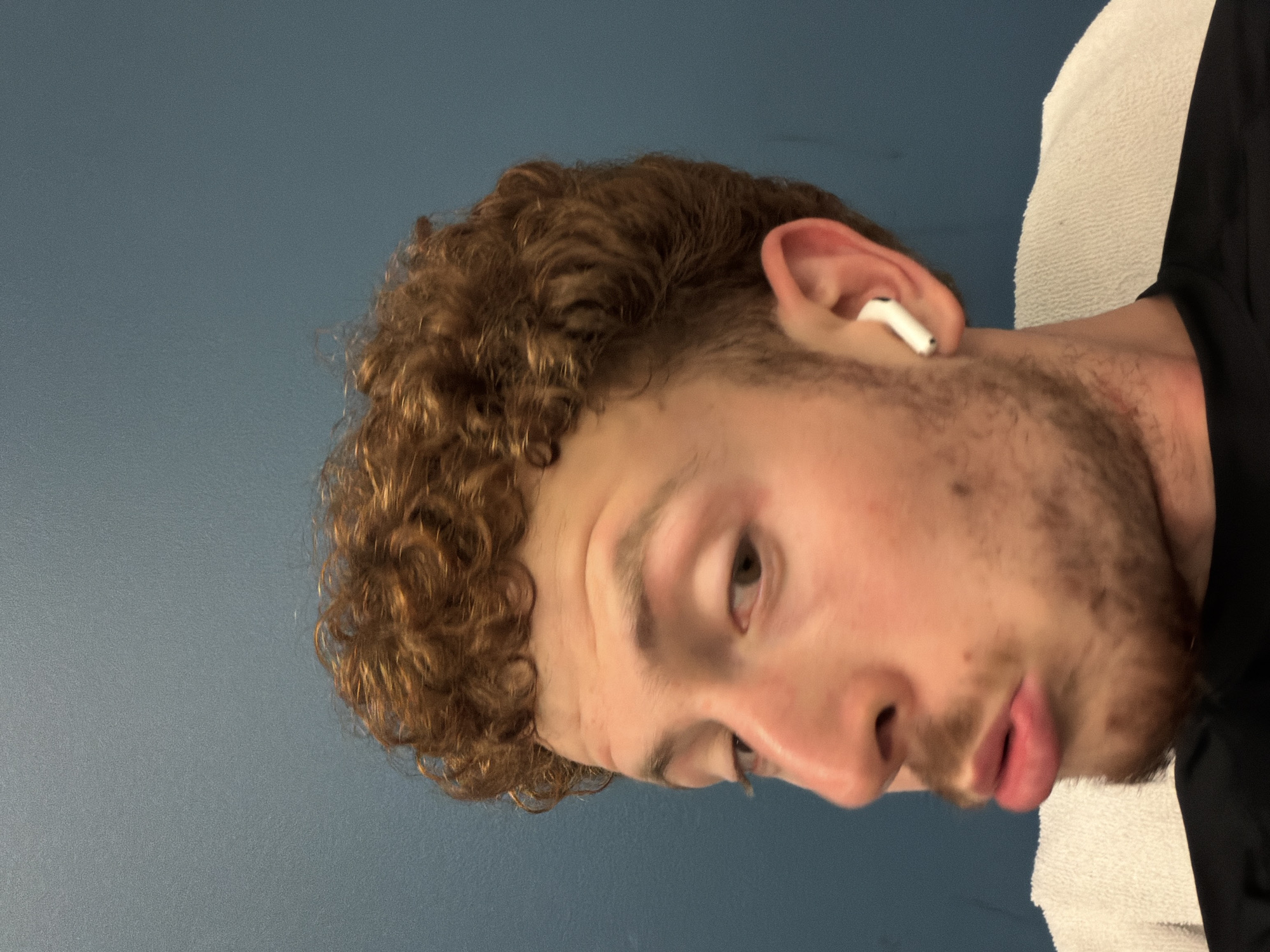 The image is a close-up, waist-up shot of a young man with curly, reddish-brown hair and a light beard, wearing a black t-shirt and a white earbud in his right ear. He is looking slightly to his left with his lips pursed. A white towel is visible draped over his shoulders. The background is a solid, dark blue wall. The image appears to be taken indoors. The location is Beachwood, United States of America.