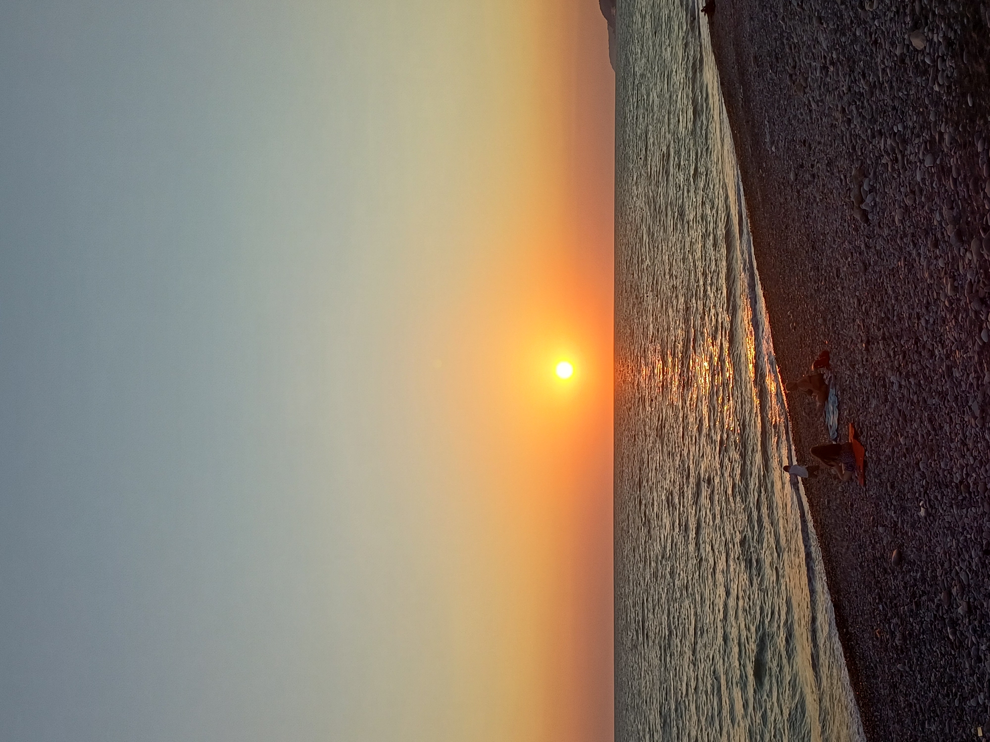 The image depicts a sunset over the sea at a pebble beach in Sochi, Russia. The sun, a bright orange orb, is low on the horizon, casting a shimmering reflection on the water. The sky transitions from a pale yellow near the horizon to a soft blue at the top. The beach is covered in dark, smooth pebbles, and a few individuals are visible in the foreground. One person is standing near the water's edge, while two others are seated on the beach, one on a blue towel and the other on an orange mat. The sea is calm with gentle waves lapping at the shore. A distant, dark landmass is visible on the right side of the frame. The overall impression is one of a peaceful evening at the seaside.