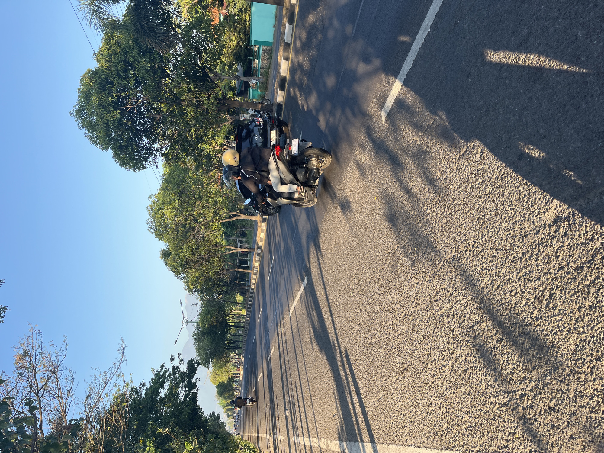 This is a photograph taken at an eye-level perspective on a street in Siring, Indonesia, during what appears to be late afternoon or early morning given the long shadows cast by the trees lining the road. The road is asphalt with white dividing lines. On the right side of the road, there is a sidewalk with a black and white striped curb, followed by a line of trees and some buildings. On the left side of the road, there are more trees and a distant mountain range. The sky is clear and blue.
In the foreground, a dark-colored sedan is partially visible behind a scooter. The scooter is carrying two people wearing helmets. The rider, on the left, is wearing a black jacket and grey pants, and the passenger on the right is wearing a yellow helmet. They are moving away from the camera. Further down the road, another person on a scooter is visible, also moving away from the camera. Several other vehicles and people are visible in the distance, suggesting moderate traffic. The overall scene depicts a typical streetscape in Siring, Indonesia, with a focus on the transportation and environment.
