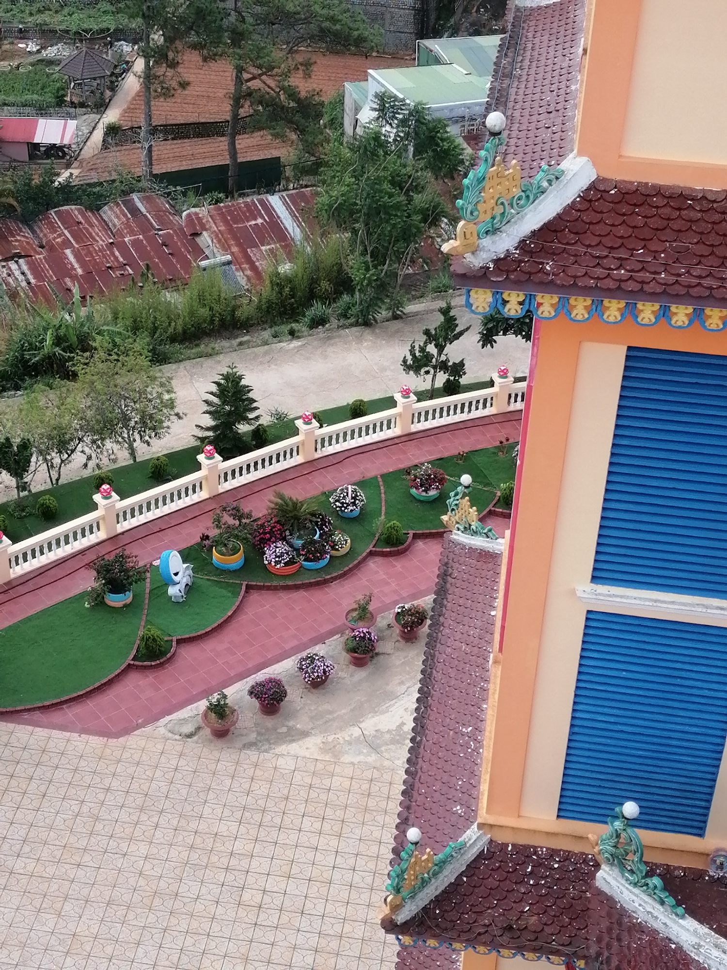 The image provides an aerial view of a temple complex in Lộc Quý, Vietnam. The foreground features the intricate, tiled roof of a temple, adorned with decorative elements in green and gold. The temple building itself is painted in light orange with large, blue-slatted windows visible. Adjacent to the temple is a paved courtyard with a repeating geometric pattern.
The middle ground showcases a well-maintained garden area with artificial green turf and red tiled pathways. Several potted plants with colorful flowers are arranged along these paths, adding vibrancy to the scene. A small, white statue of an animal is also present in the garden. A white, ornate railing encloses this garden area, separating it from a road or pathway beyond.
The background reveals a more rustic landscape, with sloping terrain covered in reddish-brown soil, likely agricultural fields. Several buildings with corrugated tin roofs, some appearing rusted, are scattered across this area. Tall green trees and dense foliage are interspersed throughout the landscape, suggesting a hilly or mountainous region. The overall impression is of a serene, spiritual site nestled within a semi-rural setting. No people are visible in the image.