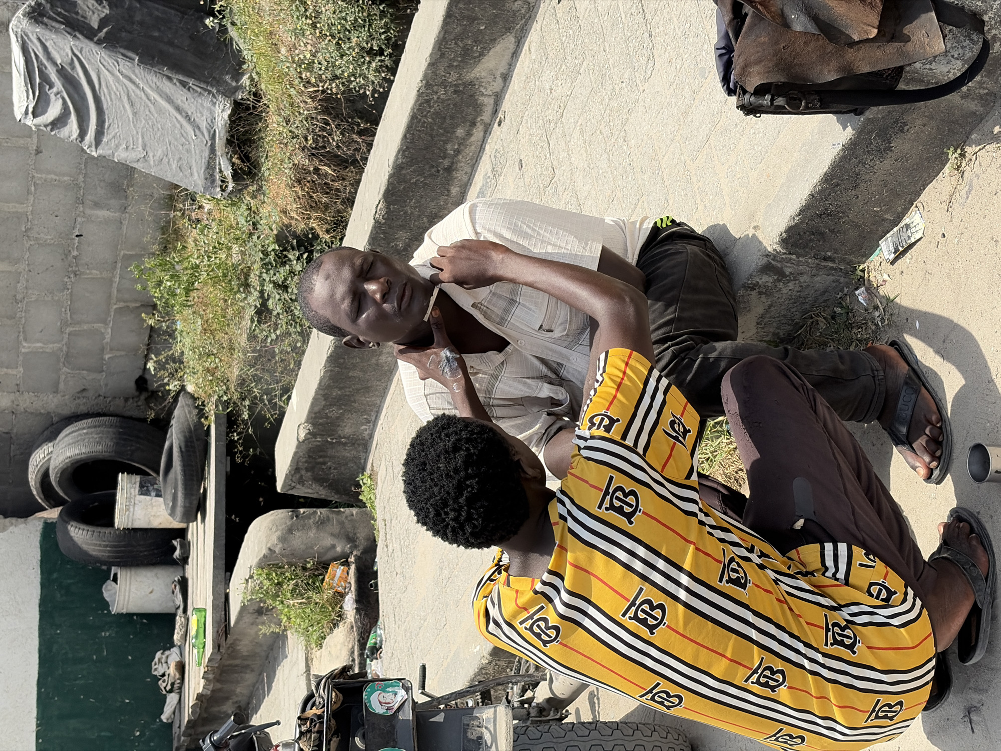 Two men are sitting on a concrete ledge in Igboefon, Nigeria. One man, wearing a yellow striped shirt, is kneeling and attending to the other man, who is wearing a light-colored shirt and dark pants. The man being attended to has a cigarette in his mouth and appears to be injured. The environment includes a stack of tires, some debris, and a cart.