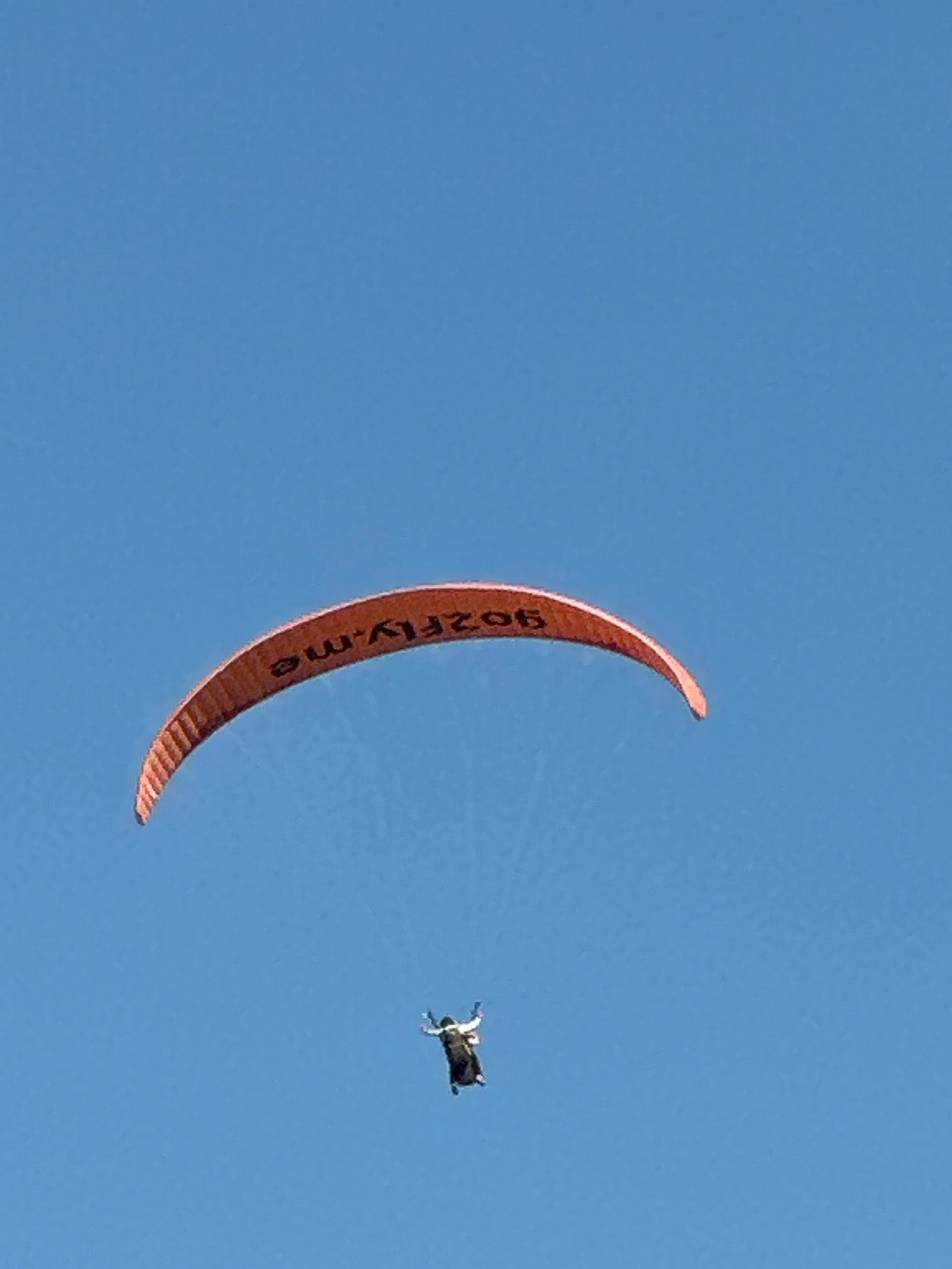 An orange paraglider with black lettering "9OZFLY.ME" is soaring through a clear blue sky. Below the paraglider, a person in a wingsuit is descending, their arms and legs extended. The scene is set against a vibrant blue sky with no discernible city or land features visible, indicating an aerial activity taking place at a high altitude. The lighting suggests daytime.