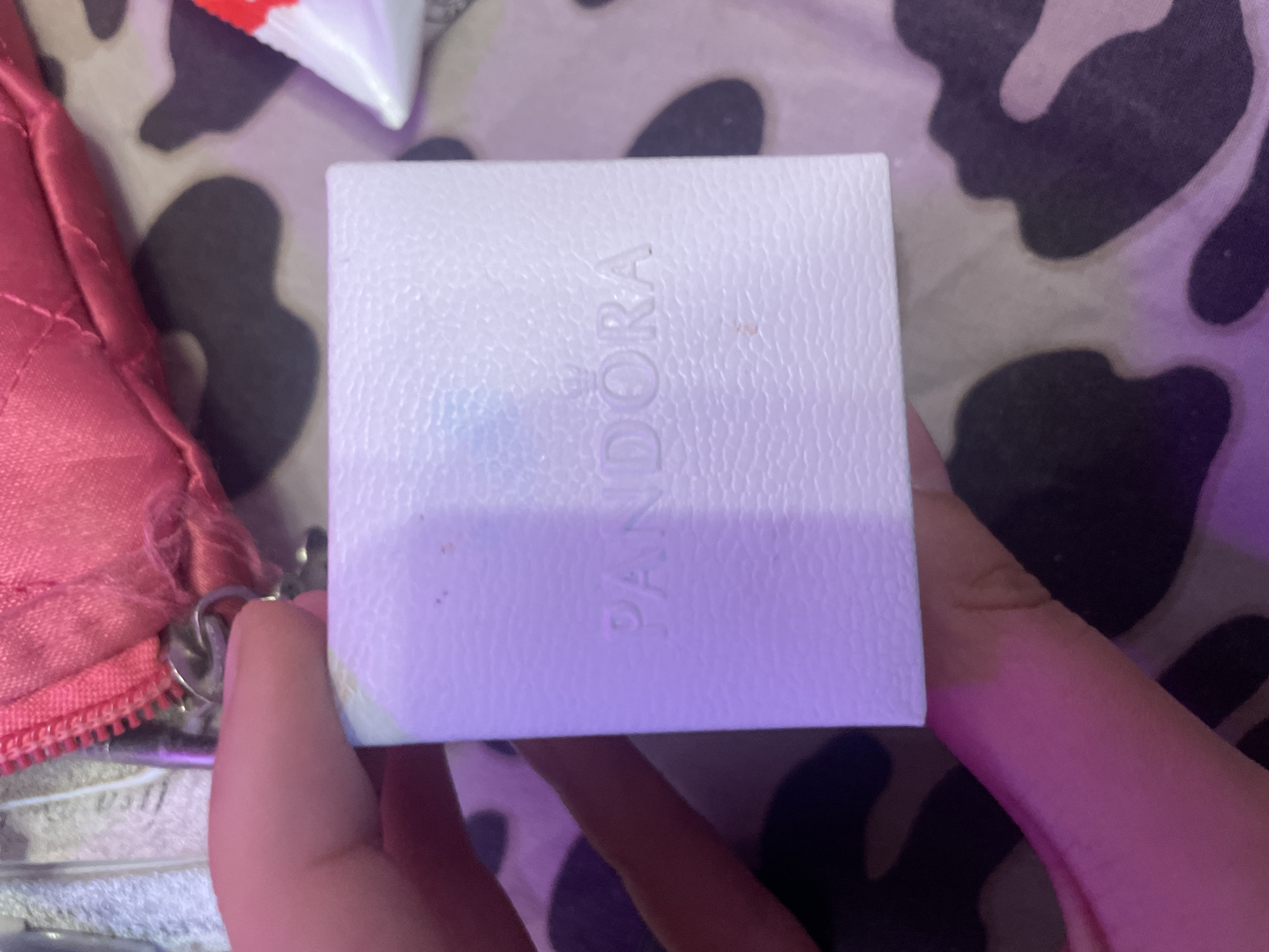 A hand holds a small, white, textured box with the embossed word "PANDORA" and a crown logo. The box is positioned in the center of the frame, against a background of what appears to be a patterned fabric with dark and light irregular shapes, possibly a leopard or cow print. In the upper left corner, a pink, quilted accessory with a zipper and a metal ring is partially visible. The overall lighting suggests an indoor setting with a purple hue cast on the scene. The location is specified as Ladozhskoe Ozero, Russia.