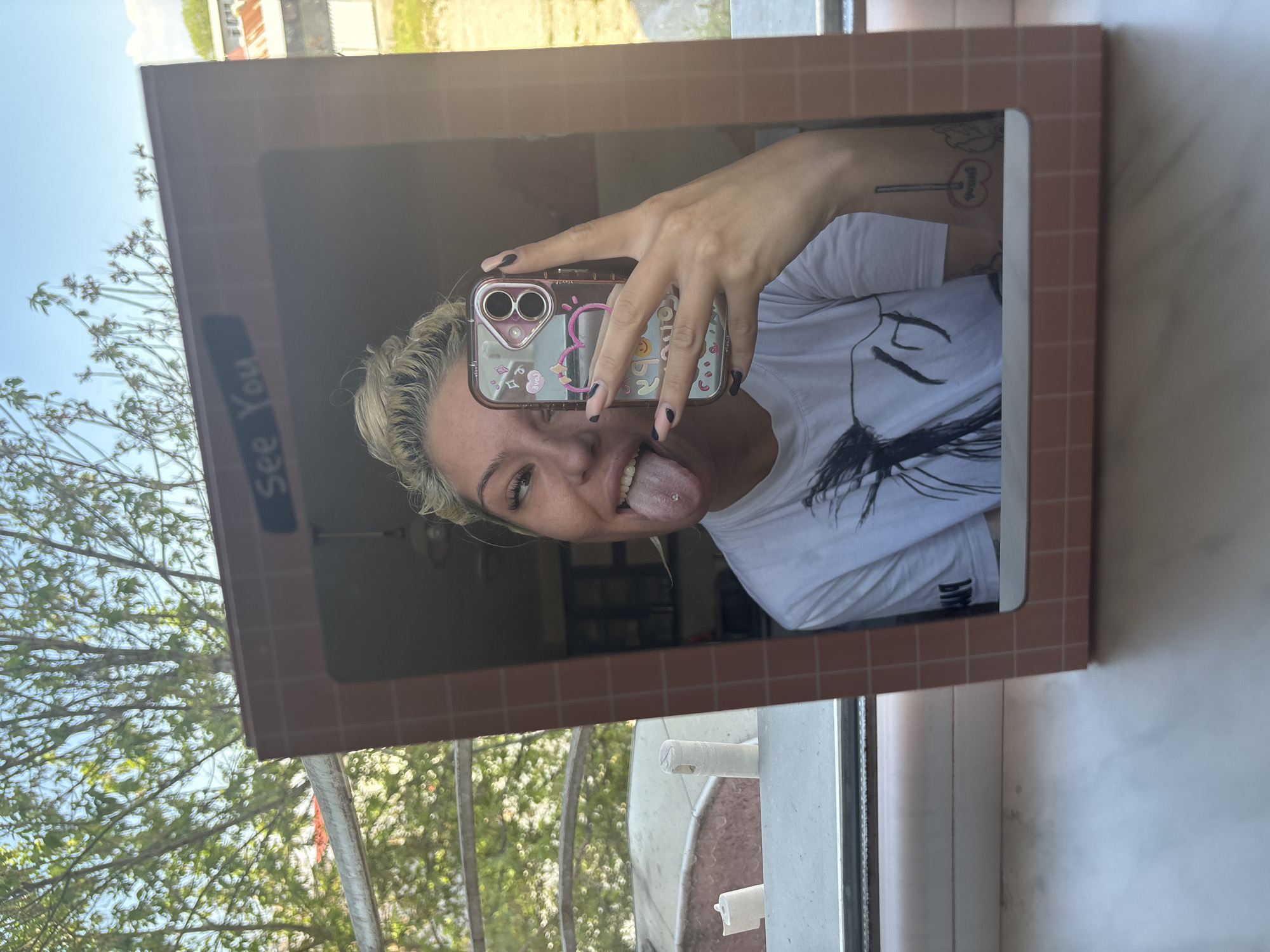 A medium shot depicts a woman reflected in a square mirror with a grid pattern frame. She is holding a smartphone with a pink and clear case up to obscure part of her face. The woman has blond hair, dark nail polish, and a tongue piercing. She is wearing a white t-shirt. The reflection reveals a room behind her. The mirror is positioned near a window with a view of trees and the sky. The location may be the city of Plovdiv, Bulgaria, based on the user's request, though this cannot be definitively confirmed from the image alone.
