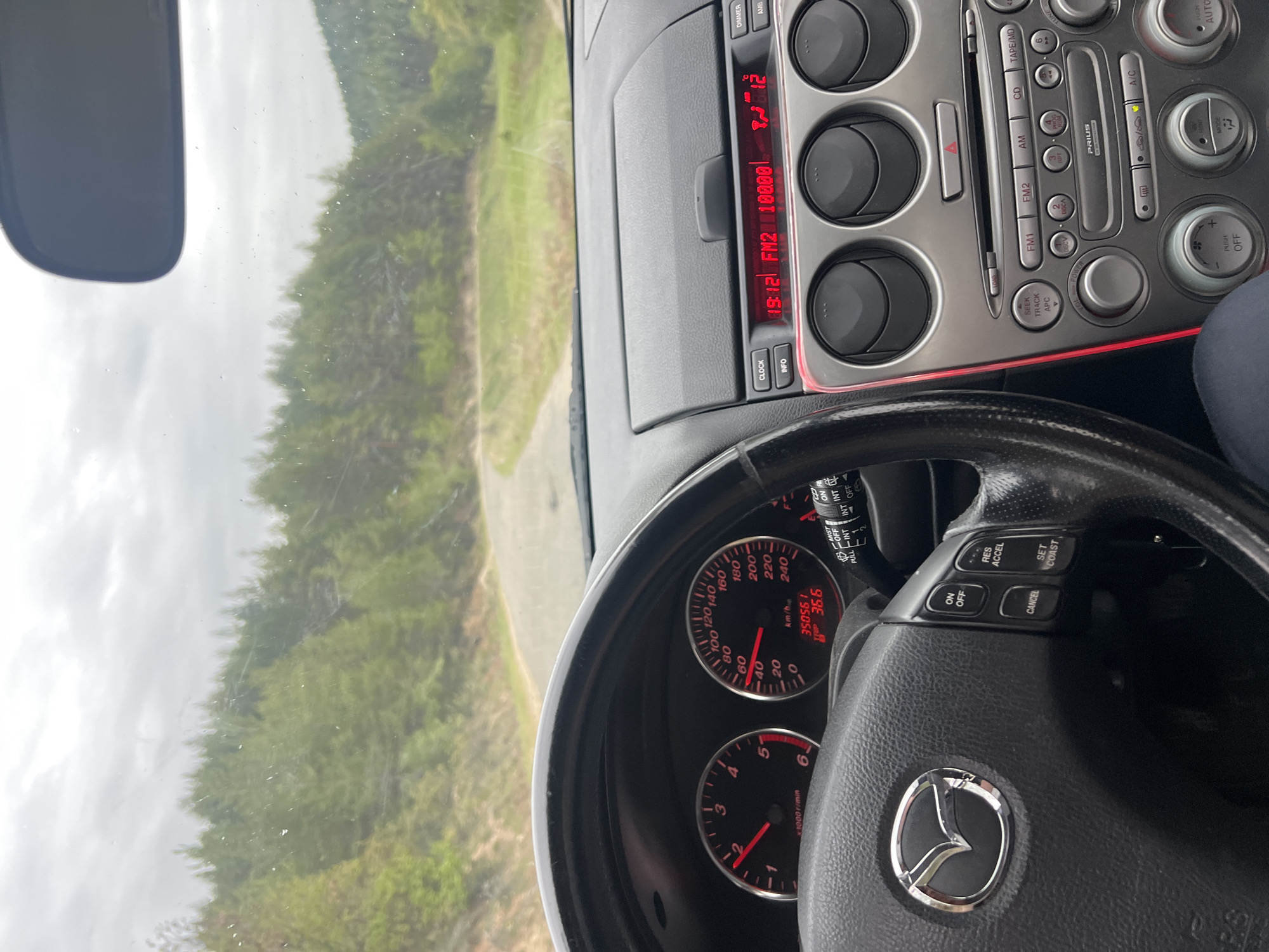 The image depicts the interior of a Mazda car while driving. The dashboard shows a mileage of 35056.1 km and a temperature of 36.6 degrees Celsius. The radio is tuned to FM2 100.00, and the time is 19:12. The driver is on a road surrounded by a forest, and the sky is overcast. The location is not in Dolno Drianovo, Bulgaria, and the information in the prompt is not contained in this image.