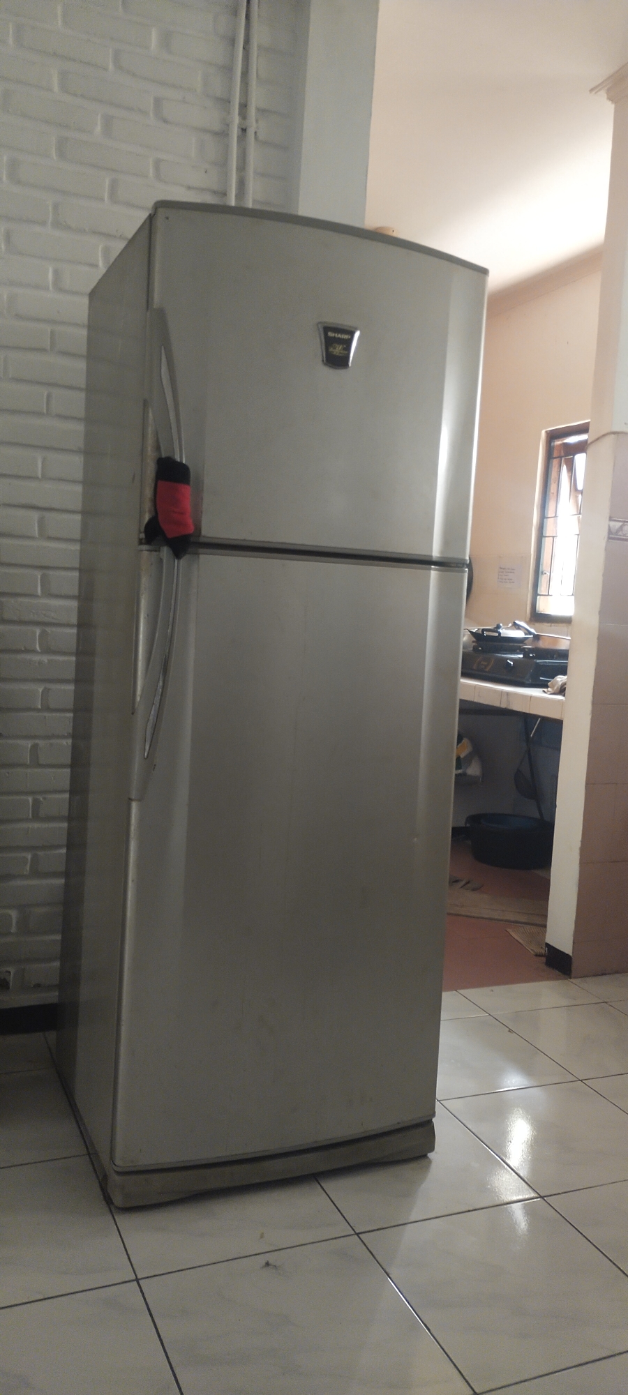 In the city of Pasirhuni, Indonesia, an indoor scene captures a top-freezer refrigerator, its metallic silver exterior reflecting the ambient light. The refrigerator is situated on a tiled floor, with the tiles appearing white and grey. To the left of the refrigerator, a white brick wall is visible, and to the right, a section of a kitchen counter with a gas stove and a window can be seen. A red and black sock is placed over the refrigerator's handle. The overall lighting suggests an indoor environment, possibly a home kitchen, with light streaming in from the window.