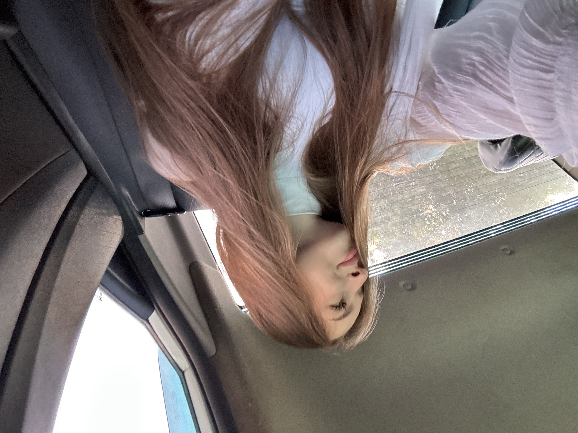 A young Caucasian woman with long, reddish-brown hair, wearing a white turtleneck shirt and a white ruffled blouse, is photographed from the chest up, inside a vehicle. She is looking to her left, with a slight smile. The interior of the car is visible, including the gray headliner, a black seatbelt, and the dark gray fabric of a car seat. Outside the car window, through the rear windshield, a blurred landscape with trees is visible. The lighting suggests an overcast day. The image appears to be a selfie taken by the subject.