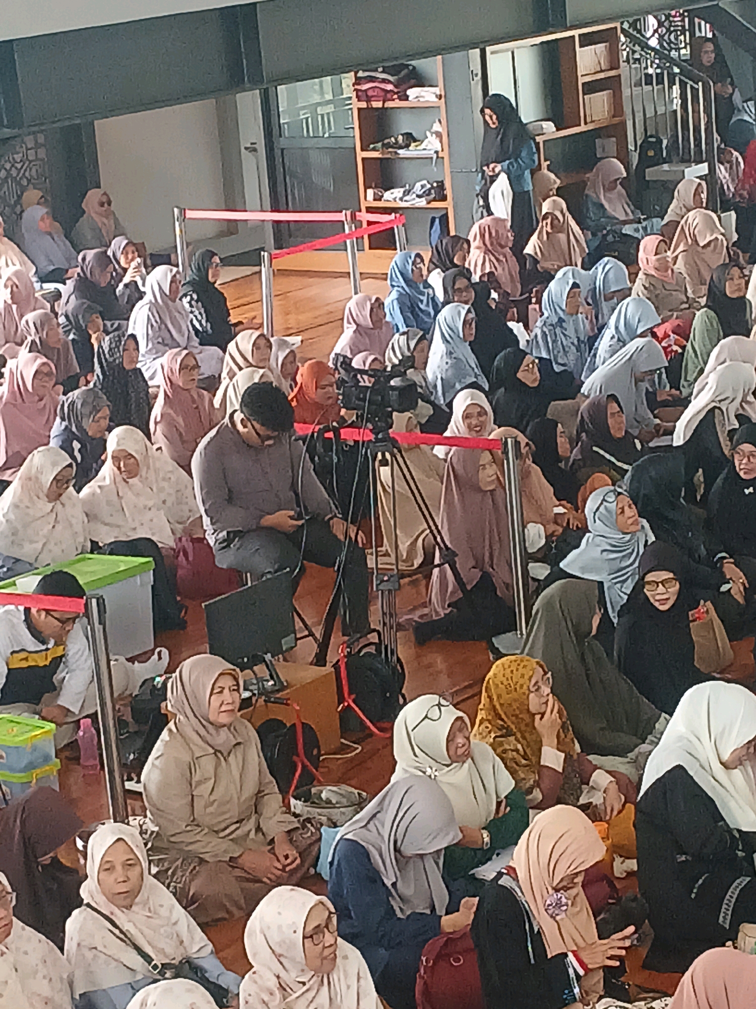 The image depicts an indoor gathering of people, predominantly women, seated on the floor. The environment appears to be a communal space, possibly a mosque or a community center in Arjasari, Indonesia, given the attire and cultural context. Many individuals are wearing hijabs, which are headscarves worn by Muslim women.
In the foreground, a man in a gray shirt and dark pants is seated, looking down at his phone. To his left, a woman in a beige hijab is also seated, with her hands clasped. Further to the left, two women in white hijabs are visible, with one looking towards the front. A green plastic storage bin is on the floor in front of them. Behind the man, a camera on a tripod is positioned, suggesting an event is being recorded or live-streamed.
The background shows more people seated, forming a dense crowd. Shelving units filled with books or other items are visible on the right side of the image, along with a staircase leading up. Red barriers are set up in the middle ground, separating areas of the space. The lighting suggests it is daytime, possibly with natural light coming from windows, though none are clearly visible. The overall atmosphere appears to be one of a seated congregation or audience.