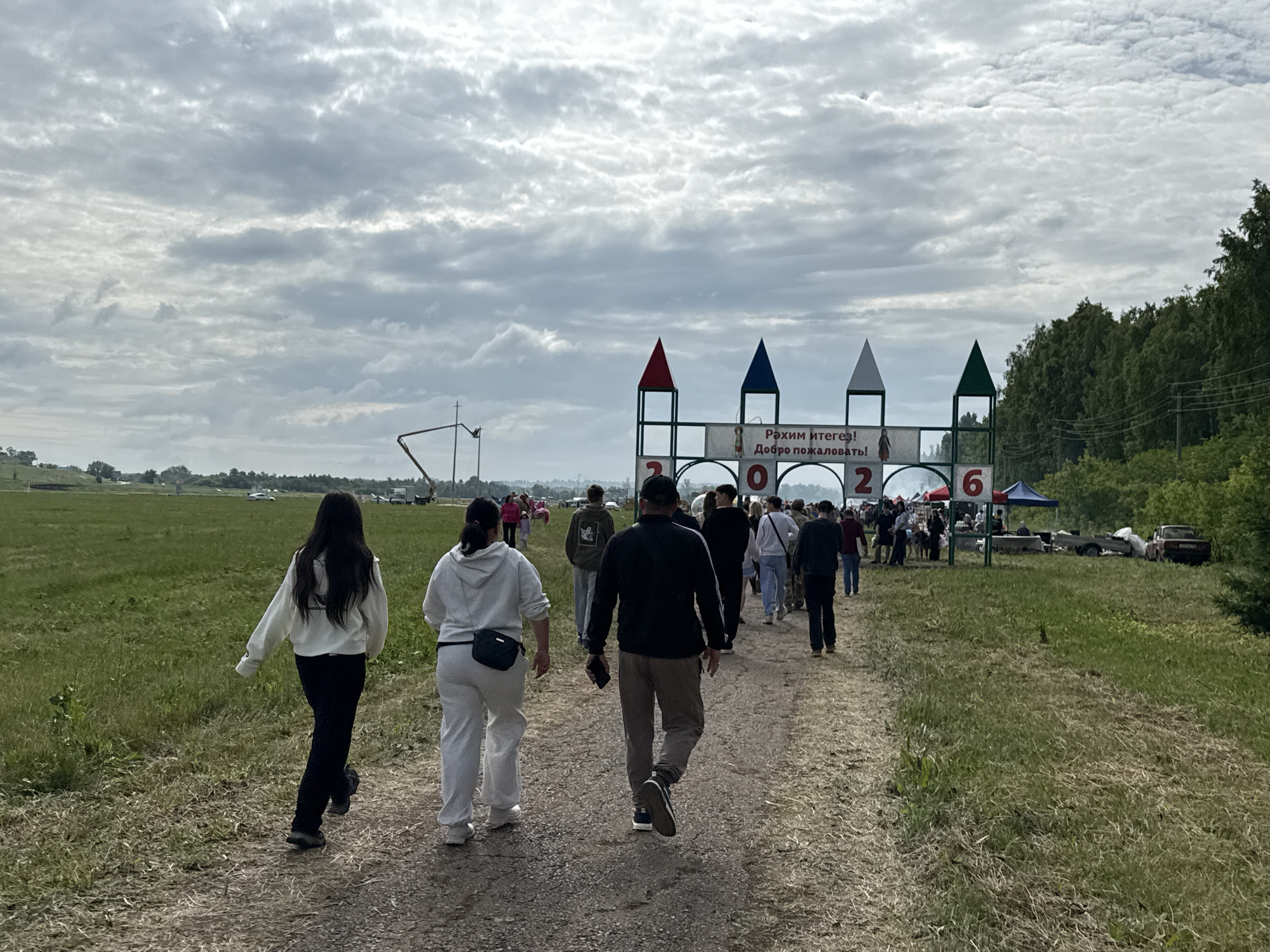 The image depicts a group of people walking along a dirt path in an open, grassy field. The sky is overcast with a dramatic cloud formation. In the background, there is a large, decorated archway with colorful triangular peaks and banners that appear to have text in Cyrillic. There are also some trees visible to the right of the archway, and more trees and fields in the distance. The scene suggests an outdoor event or gathering. The presence of the archway with Russian text indicates it is likely located in Russia. Based on the provided context, the location is identified as Старое Субханкулово, Russia.