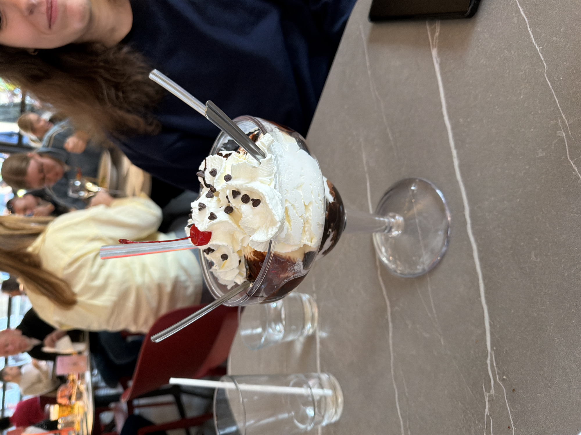 The image displays a delectable ice cream sundae as its central subject, resting on a gray, mottled table with distinct white vein-like patterns. The sundae is served in a tall, clear glass dish, filled with layers of light-colored ice cream (likely vanilla), rich chocolate sauce, and generously topped with whipped cream and chocolate chips. A bright red maraschino cherry and a red plastic straw garnish the dessert, alongside two metallic spoons or spatulas inserted into it.
To the right of the sundae, two empty clear drinking glasses are visible on the table, one containing a white straw. Part of a black smartphone is also visible on the upper right edge of the table.
In the upper left of the frame, the face and upper torso of a person with long, wavy brown hair are partially visible, looking towards the sundae with a slight smile. The background is blurred, showing other patrons seated at tables in what appears to be an indoor cafe or restaurant. The presence of multiple people and dining settings suggests a casual and social environment. Parts of red chairs are also discernible in the mid-ground. The scene depicts a relaxed moment of enjoying a dessert, possibly in Milan, Italy, as per context, but without specific cues in the image itself. No visible text is present.