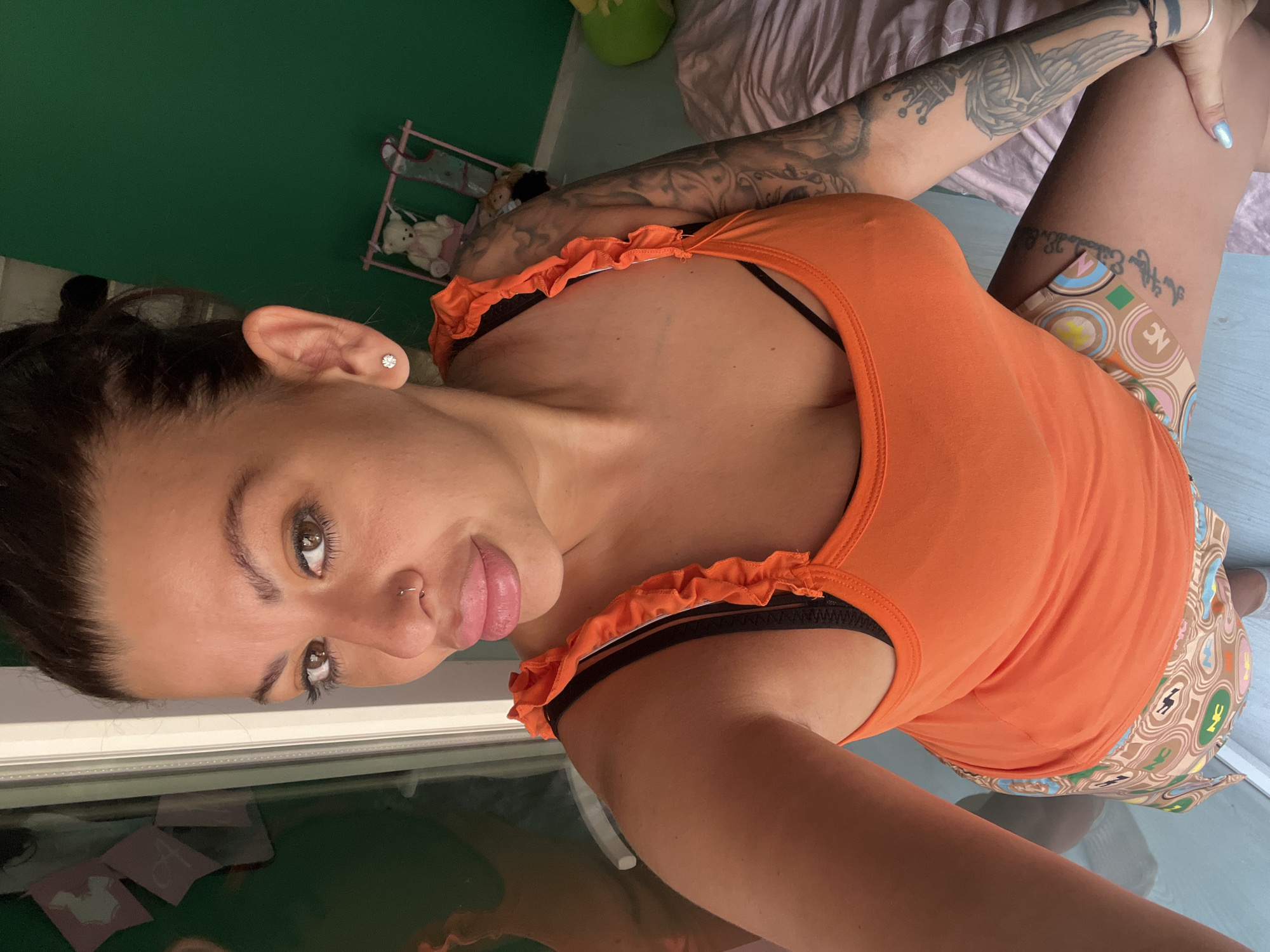 The image shows a woman with dark hair pulled back, wearing an orange tank top with ruffled straps and patterned shorts. She has a nose ring and wears a diamond stud earring. Visible tattoos adorn her arms and thigh. She is positioned indoors, with a window to her left reflecting a green wall and some decorative items, including a small teddy bear on a pink swing. The background also includes a bed with a pink comforter. The scene appears to be a domestic setting. No elements in the image suggest a specific location within the city of Plovdiv, Bulgaria.