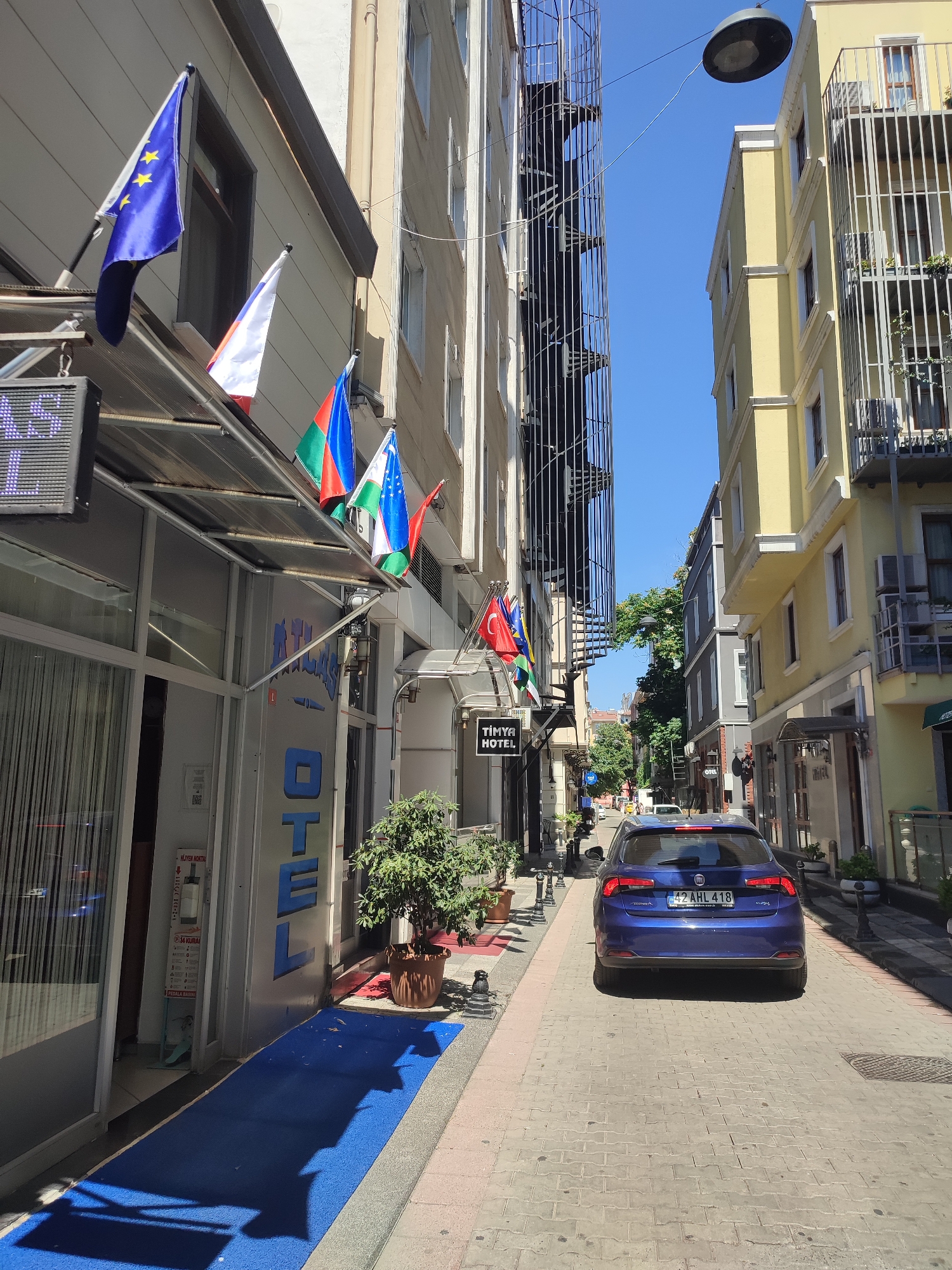 The image depicts a narrow street in Kadıköy, Turkey, on a bright, sunny day. The street is lined with buildings of varying heights and architectural styles, including hotels and apartments. On the left side of the street, there is a hotel with several flags displayed, including the European Union flag, the Russian flag, and what appear to be other national flags. A blue carpet leads to the entrance of the "TİMYA HOTEL." A blue Fiat car is parked in the middle of the street, facing away from the camera. The street is paved with cobblestones, and there are potted plants and decorative lampposts along the sidewalks. The sky is clear and blue.