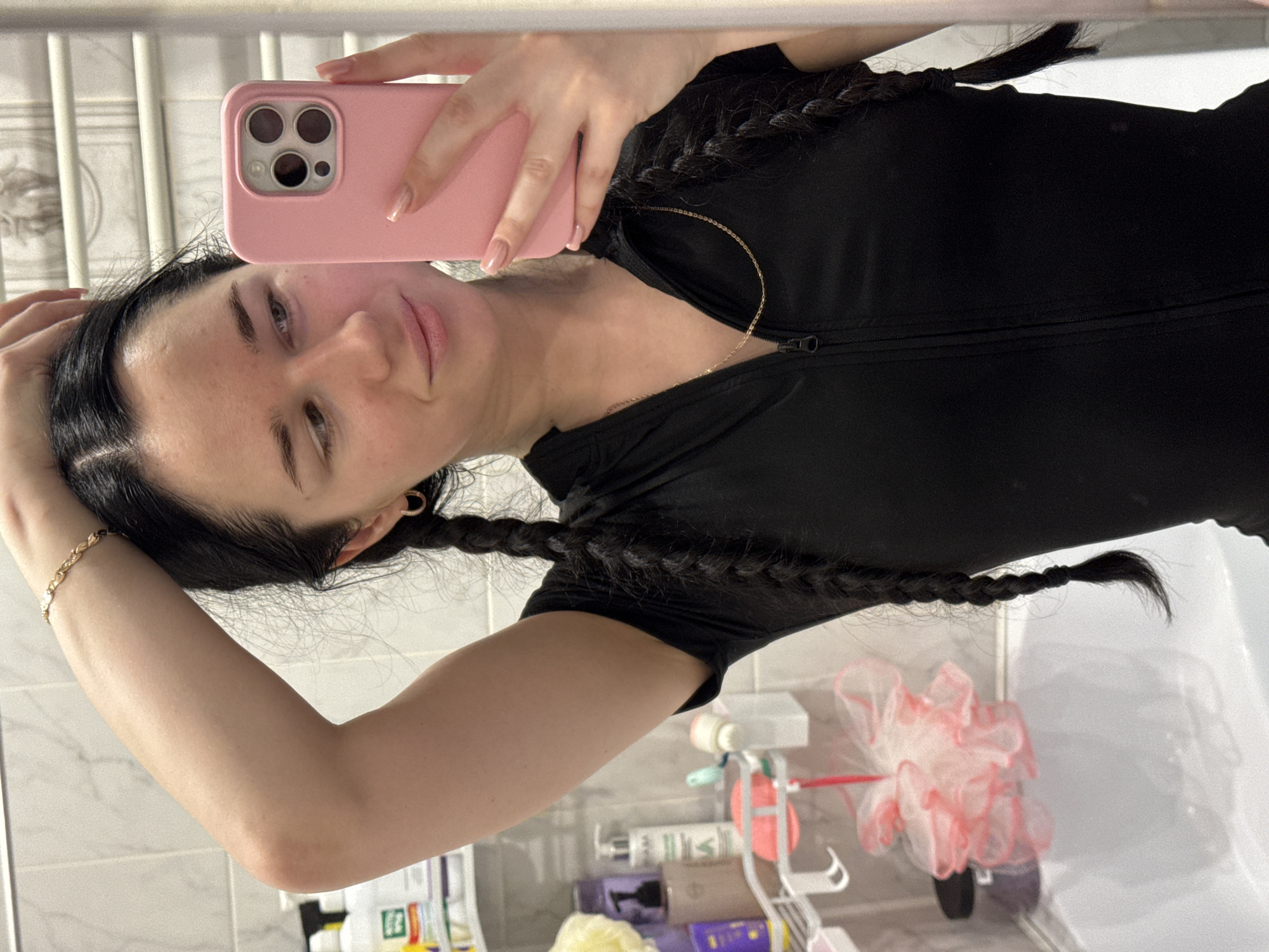The image shows a young woman with dark hair styled in two braids, wearing a black zippered top. She is holding a pink smartphone in her right hand, taking a selfie in a bathroom mirror. Her left arm is raised, touching her head. A gold bracelet is visible on her left wrist, and a delicate gold necklace is worn around her neck. Behind her, a bathroom shelf is stocked with various toiletries, and a pink loofah hangs near a bathtub. The tiles on the wall are white. The scene is indoors, likely in a residential bathroom. There is no indication that this location is in Novosibirsk, Russia, or any specific city.