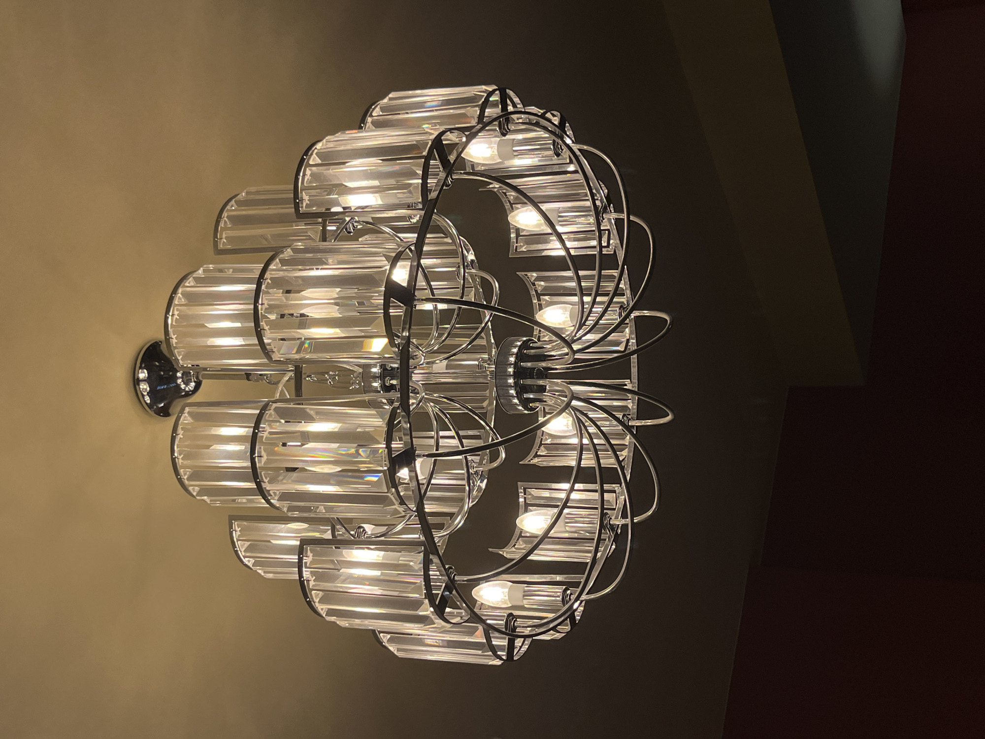 A multi-tiered crystal and metal chandelier is centrally depicted, suspended from a light beige ceiling in an indoor location within Bečići, Montenegro. The chandelier features a polished metal framework, likely chrome, supporting numerous vertical, rectangular, faceted clear crystal elements arranged in two main tiers. The upper tier consists of crystal panels, while the lower tier incorporates multiple exposed, lit, candle-shaped incandescent bulbs, casting warm light and producing prismatic refractions within the crystal components. The ceiling is uniformly light beige. Below the chandelier, the background transitions to a darker, shadowed wall, with a distinct horizontal line suggesting a material or color change. A section of a dark red wall is visible at the bottom left quadrant of the frame. No individuals are present.