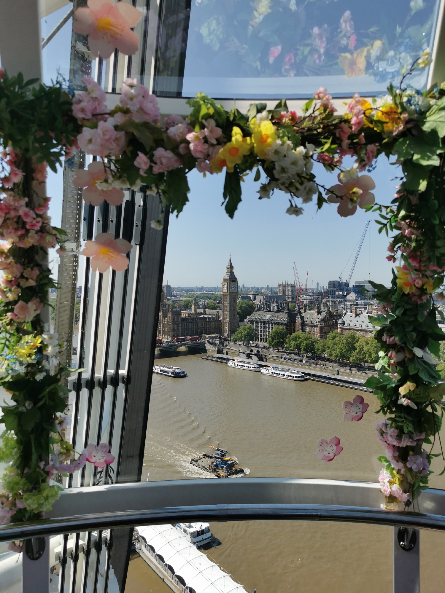 The image presents a view of London landmarks from a high vantage point, possibly within the London Eye. In the distance, Big Ben and the Houses of Parliament are clearly visible alongside a number of cranes. The River Thames flows in the foreground, carrying several boats. The scene is framed by a floral garland with pink and yellow flowers, which adorns the structure in which the photo is taken. The day appears sunny and clear, casting light across the city.