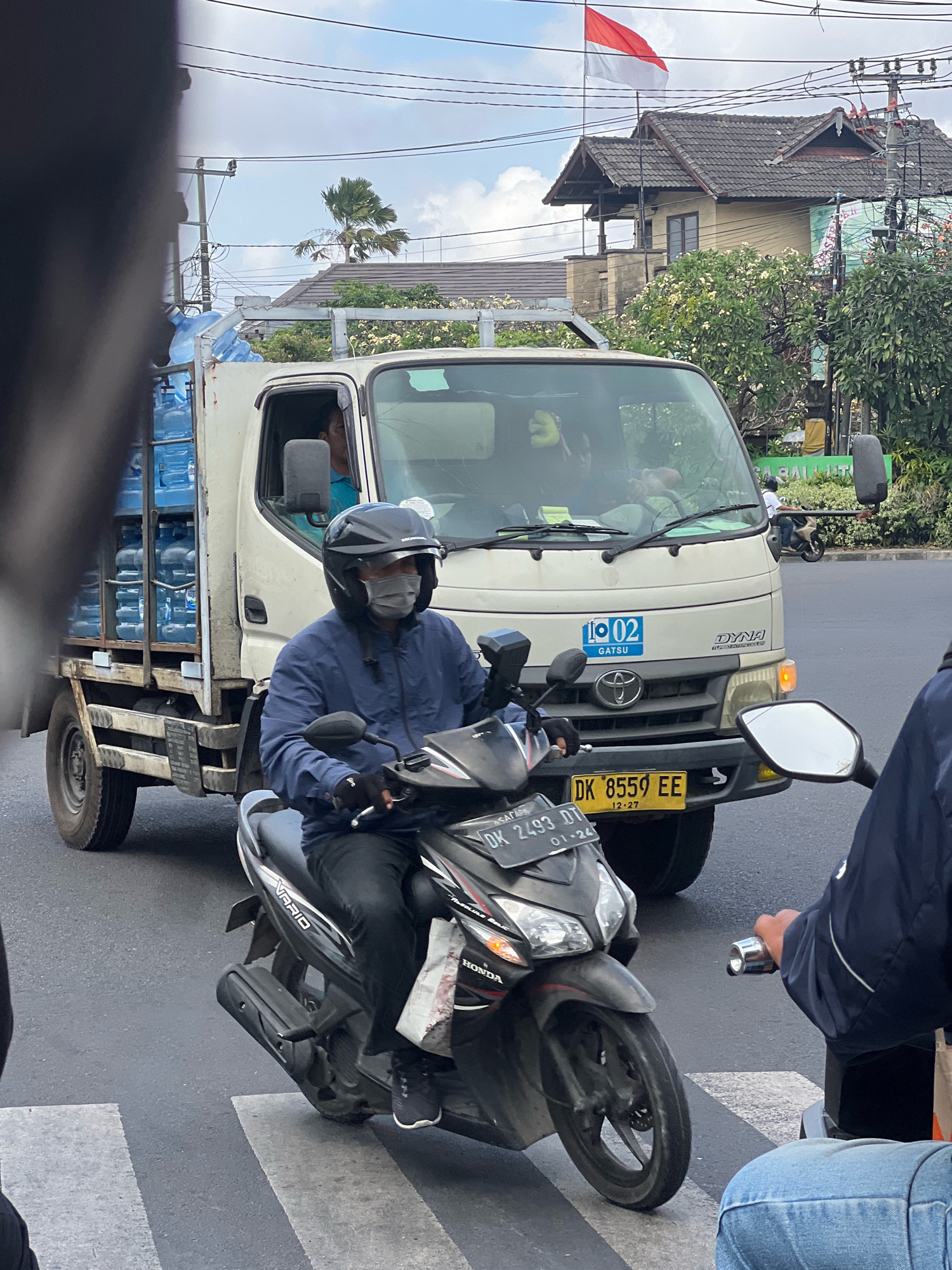 In Denpasar, Indonesia, a delivery truck loaded with blue water jugs is stopped at a crosswalk. The truck, a white Toyota Dyna, has a license plate starting with "DK." A motorcyclist wearing a blue jacket and a black helmet is in the foreground, also stopped at the crosswalk. The motorcyclist's scooter has a license plate reading "DK 2493 DT." Another person on a motorcycle, wearing a blue jacket, is partially visible to the right. In the background, a red and white Indonesian flag flies from a building. The sky is partly cloudy, and palm trees and other greenery are visible. The scene depicts a typical street intersection in Denpasar.