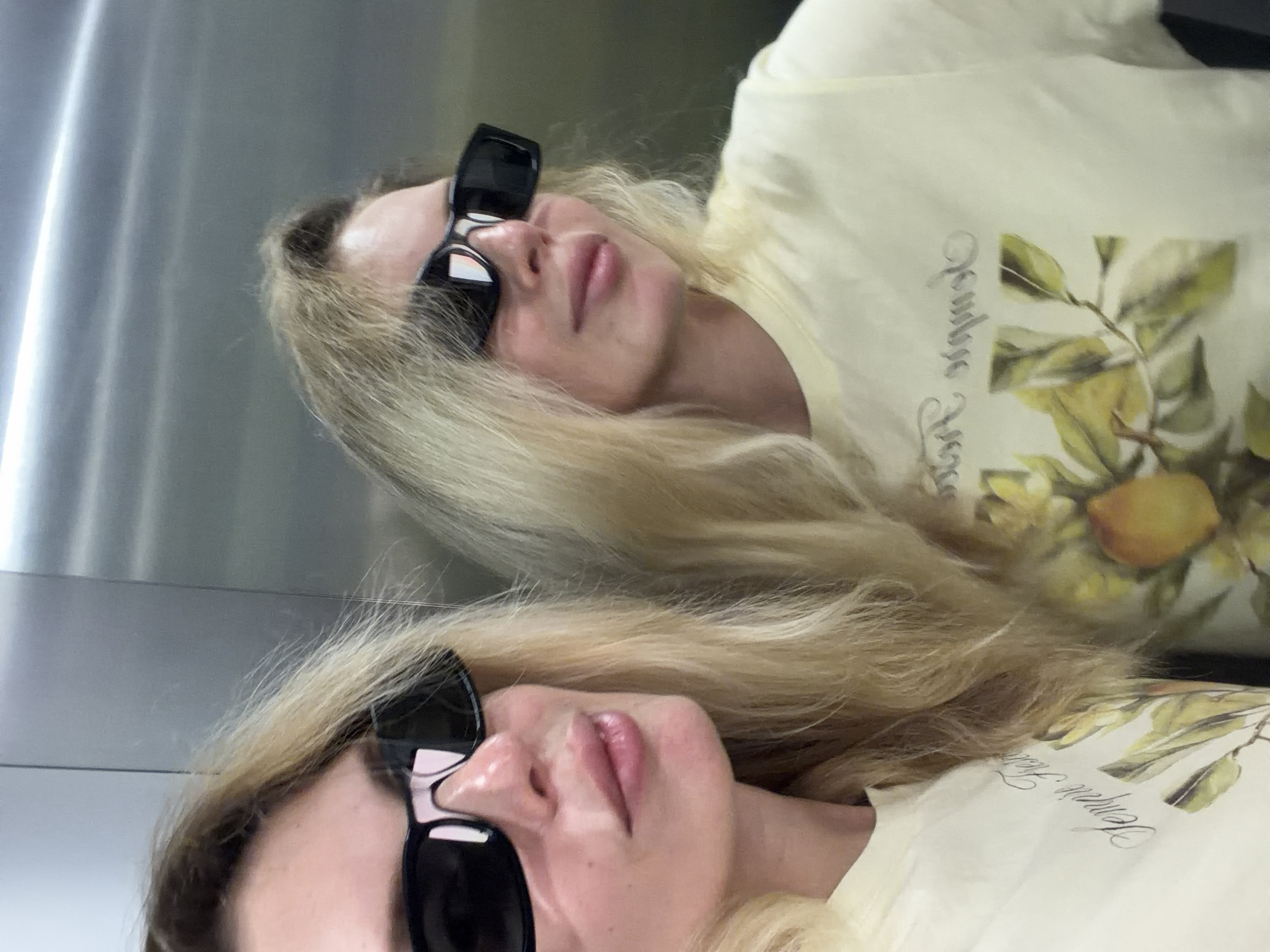 A female individual with long blonde hair, wearing dark, rectangular sunglasses, is captured in a mirror selfie within what appears to be an elevator. She is wearing a light yellow short-sleeved t-shirt featuring a graphic print of a lemon or citrus branch with green leaves and a stylized text inscription. The individual's reflection is prominently visible to the right, showing the same attire and features. The background consists of polished metallic panels, characteristic of elevator interiors, with horizontal seams or divisions visible. Her lips are slightly parted, revealing light lip color. There are no discernible visual cues within the image to indicate its specific location within the city of Budva, Montenegro.