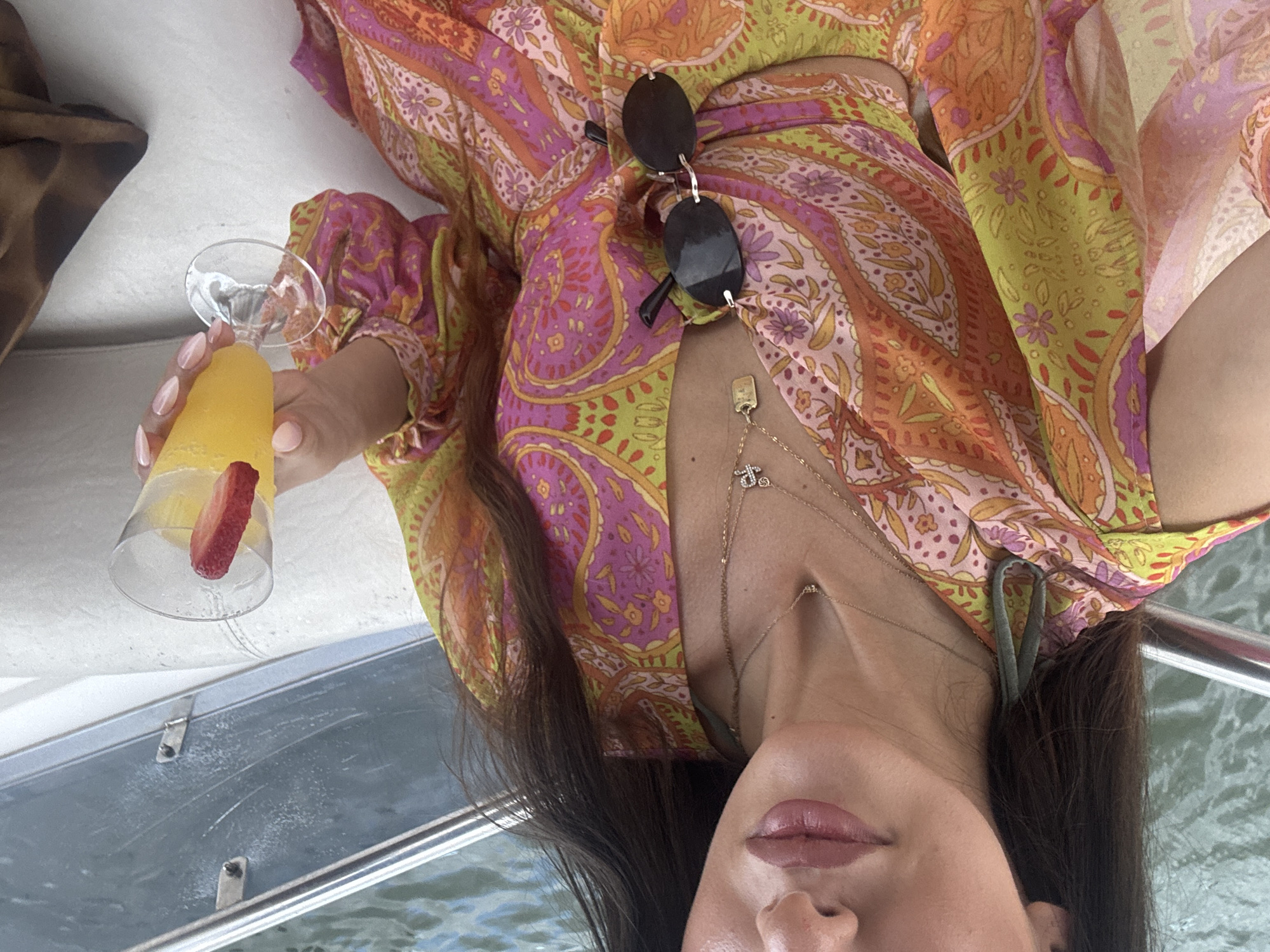 A woman is lying on a boat with a drink in her hand. The woman has dark hair and is wearing a colorful patterned dress. The drink is a cocktail with a strawberry garnish. There are sunglasses resting on her dress. The boat appears to be on the water, possibly in Miami, given the presence of water and the overall aesthetic.