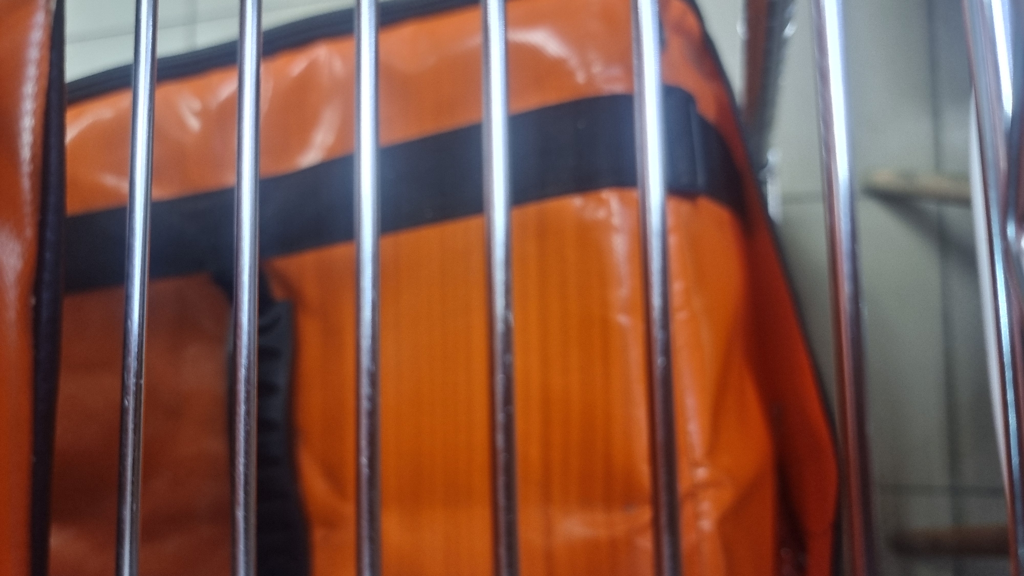 The image shows a close-up of an orange and black bag with metal bars in front of it. The bag appears to be made of a shiny, possibly vinyl or synthetic material, and has a pattern of horizontal orange sections separated by black stripes. The metal bars are thin and reflective, and are positioned horizontally across the frame, obscuring parts of the bag. The background is out of focus and appears to be a light-colored, possibly a wall or a partition. There are no people or specific identifiable landmarks visible in the image that would allow for determination of its location within the city of Megion, Russia. The image is a still shot and captures a static scene.
