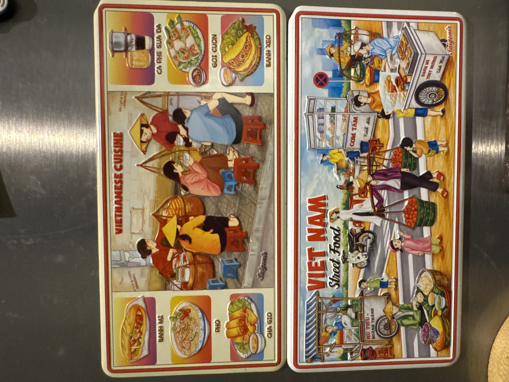 Two rectangular plates, side by side, showcase Vietnamese cuisine and street food in Saigon. The plate on the left is titled "Vietnamese Cuisine" and features illustrations of common Vietnamese dishes, including "Ca Phe Sua Da," "Goi Cuon," "Banh Xeo," "Banh Mi," "Pho," and "Cha Gio." Illustrations of people sitting on small stools eating are also present. The plate on the right is titled "Viet Nam Street Food" and depicts a lively street scene with various food vendors and people, including vendors selling "Com Tam," "Banh Mi Thit Nuong," and "Hu Tieu Hoanh Thanh." The plates are designed to resemble embossed artwork, with raised elements. The plates are sitting on a grey surface. The city of Kuta Selatan, Indonesia is not visible in the image.