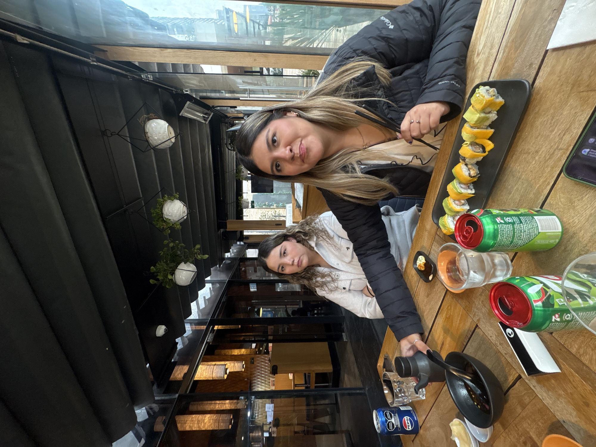 The image depicts two women at a table in what appears to be a restaurant, potentially in Mexico City, Mexico. The setting features elements of an enclosed terrace or a restaurant with a glass ceiling, allowing natural light.
One woman is wearing a black jacket and the other is wearing a gray or white shirt with a black jacket as well. They are seated at a wooden table.
On the table, there is a plate of sushi rolls, two cans of what appear to be soft drinks, several glasses, and a small black bowl.
The background of the image shows a view of the city with buildings and trees, suggesting the restaurant is located in an urban area with some greenery. The interior design of the restaurant is contemporary, with hanging light fixtures and a mixture of glass and metal structures.