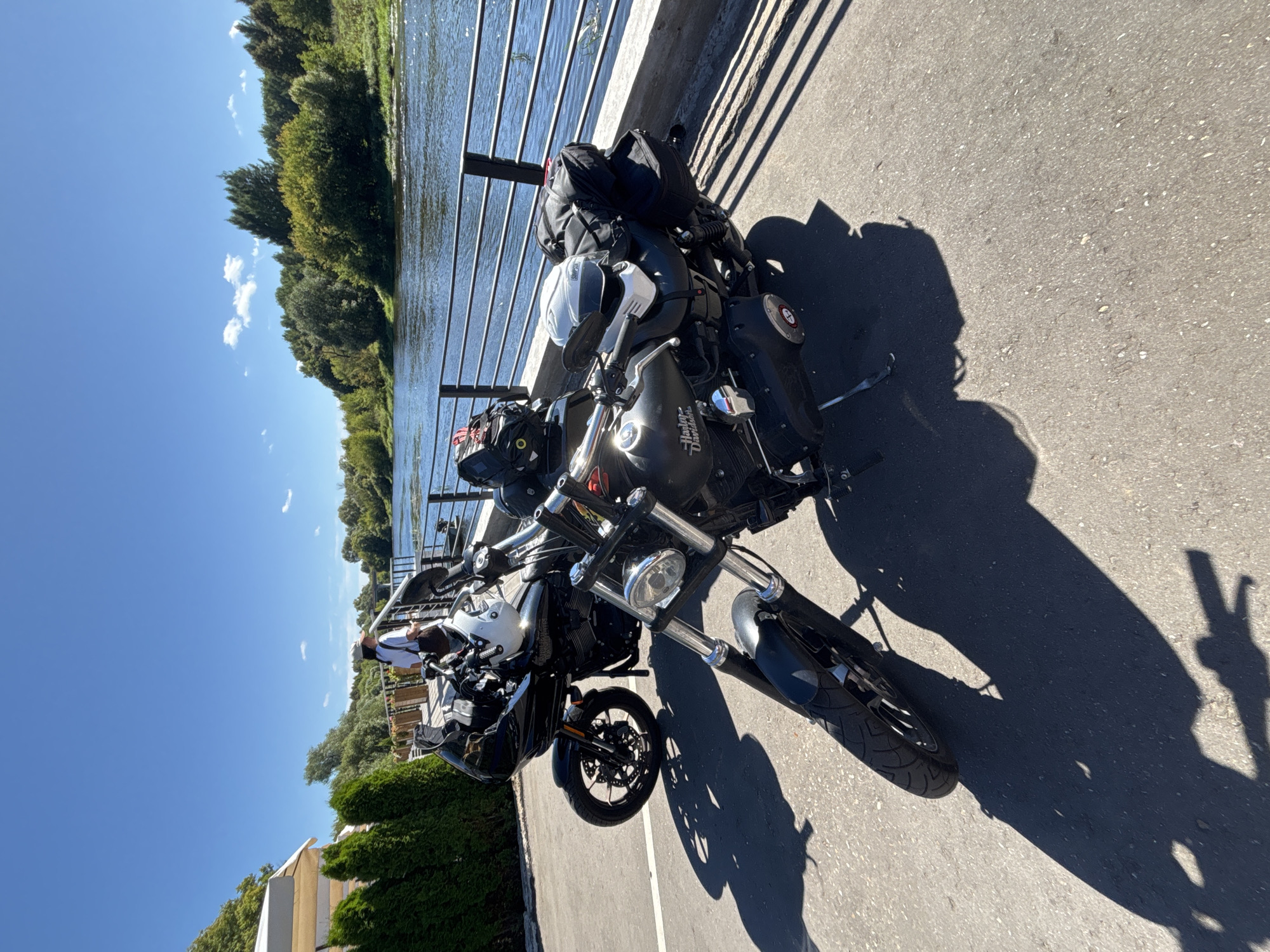 Two black Harley-Davidson motorcycles are parked on an asphalt surface next to a river. The motorcycle in the foreground is closer to the camera, with its front wheel and handlebars visible. It is equipped with saddlebags and a helmet on the handlebars. The second motorcycle is parked behind the first, with its front fairing and handlebars visible. A person wearing a white shirt and a cap stands on a metal railing overlooking the river, facing away from the motorcycles. The river is wide and appears calm, with trees and greenery lining its banks. The sky is clear and blue with a few scattered clouds. The scene suggests a stop at a scenic location, possibly a park or a riverside cafe, in the city of Zvenigorod, Russia.