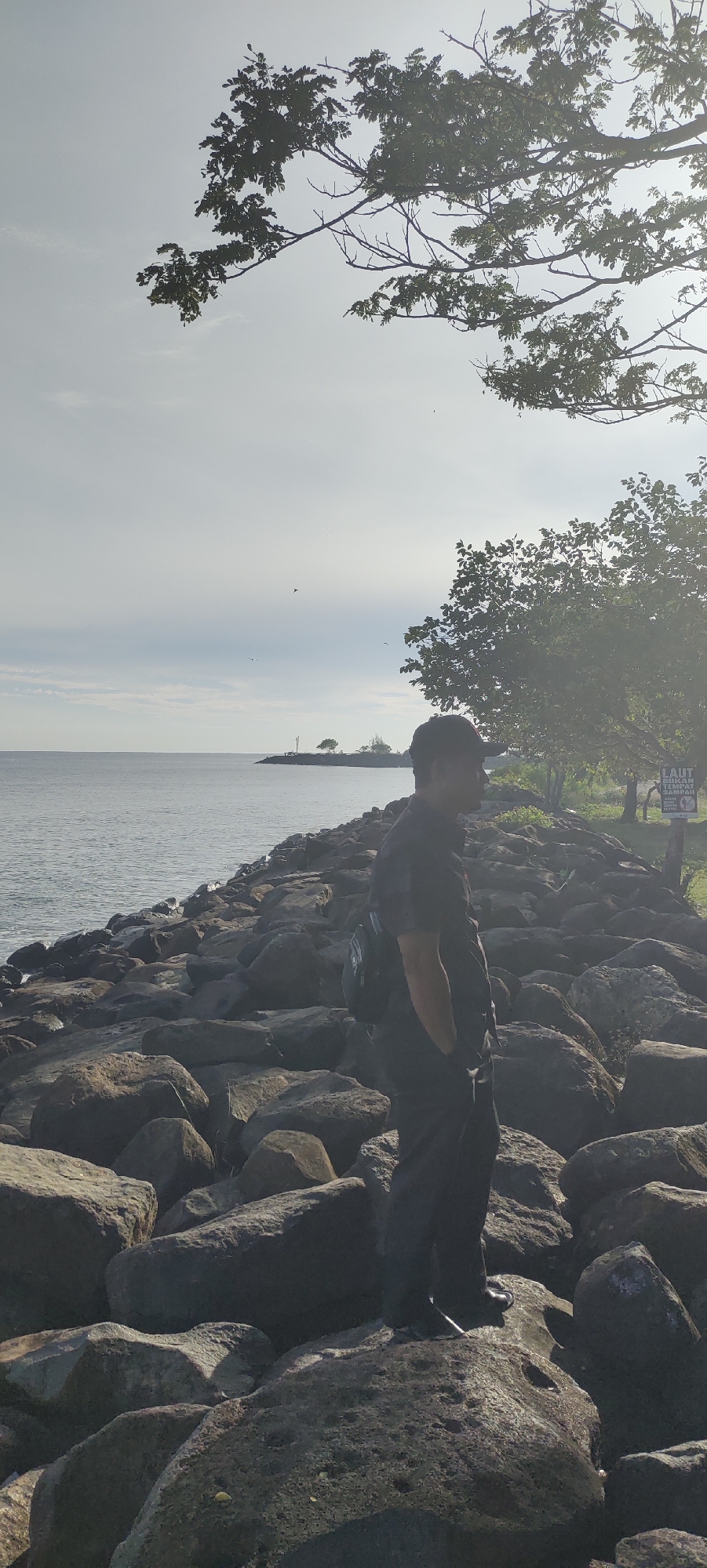 The image depicts a man standing on a rocky shoreline, looking out at the sea. He is wearing a black shirt and pants, and a backpack. The sea is calm, and the sky is overcast. There are trees in the background, and a sign that reads "LAUT BUKAN TEMPAT SAMPAH" which translates to "THE SEA IS NOT A TRASH DUMP". The scene is likely located in Banda Aceh, Indonesia, as suggested by the Indonesian text on the sign and the tropical vegetation. The rocky shoreline suggests a coastal area, possibly a harbor or a beach breakwater. The overall mood of the image is peaceful and contemplative.