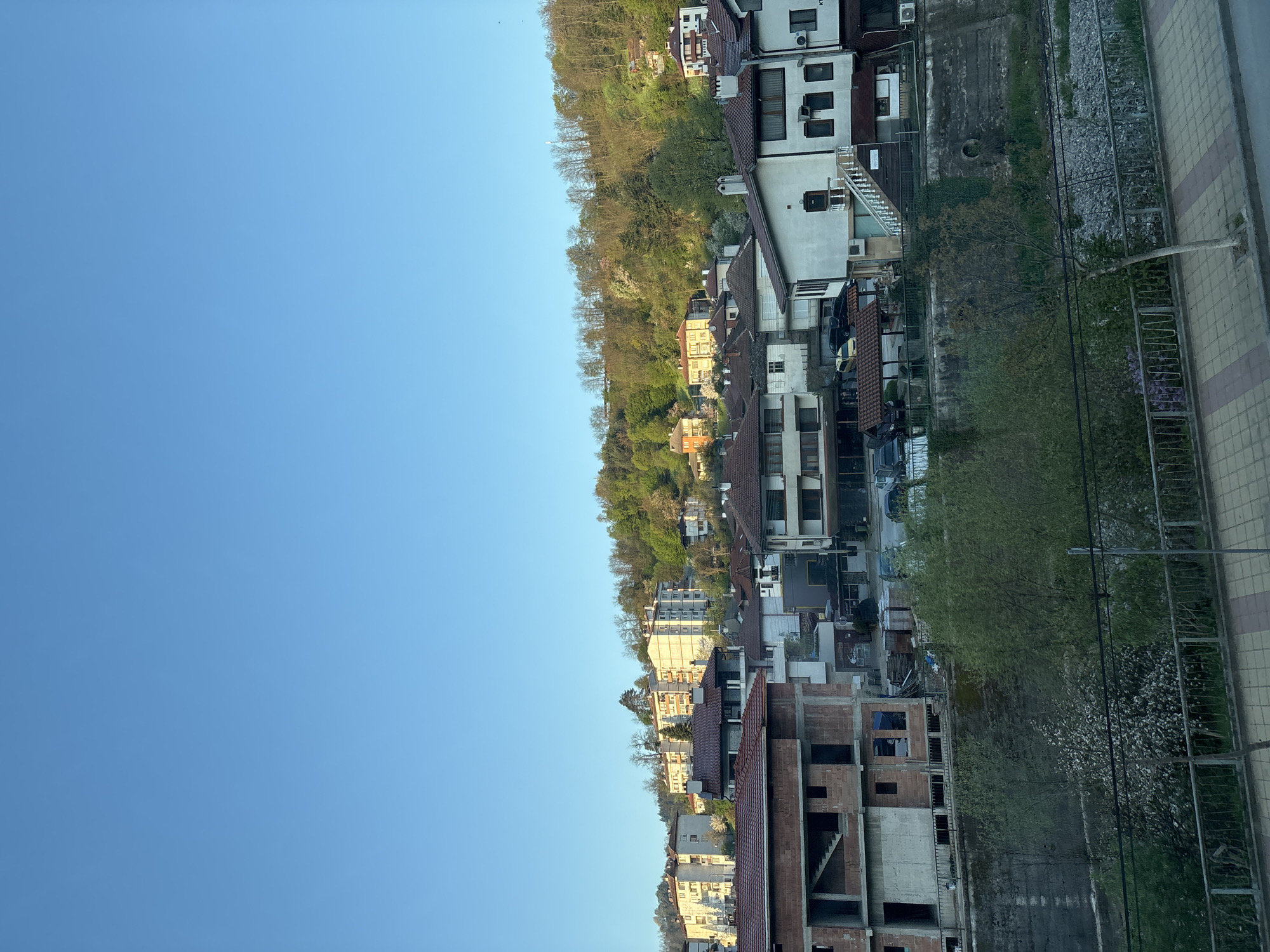 The image depicts a panoramic view of a town situated at the base of a forested hill under a clear, bright blue sky.
In the upper portion of the frame, an expansive, unblemished blue sky dominates, gradually lightening towards the horizon line. Below the sky, the landscape unfolds with a blend of natural and urban elements. A densely wooded hill, covered in a mix of verdant green trees and some with bare branches, rises prominently, creating a natural backdrop for the settlement.
The town itself features a collection of buildings, primarily residential, varying in architectural style and age. Many structures have reddish-brown tiled roofs, while their facades range from white to cream and pale yellow. Balconies, windows, and outdoor air conditioning units are visible on several buildings. Towards the lower left, a multi-story building is conspicuously under construction, with exposed brickwork and concrete frames, indicating ongoing development.
In the foreground, running along the bottom right of the image, is a paved sidewalk characterized by a repeating pattern of light and dark square tiles. A metal fence separates the sidewalk from a strip of unkempt ground below, which consists of rocks, dirt, and sparse vegetation, potentially a dry riverbed or a drainage area. Electrical wires are also visible spanning across this foreground section. Closer to the ground, scattered trees display white blossoms, suggesting it is springtime.
The scene portrays a quiet, everyday moment in a town, with no specific human activity visible. The clear sky and bright illumination suggest a sunny day, possibly late morning or early afternoon, with crisp details. The overall atmosphere is tranquil. No legible text is visible on any of the buildings or signs. Given the location context, this is a typical scene from Zlatograd, Bulgaria, showcasing its blend of natural beauty and urban life.