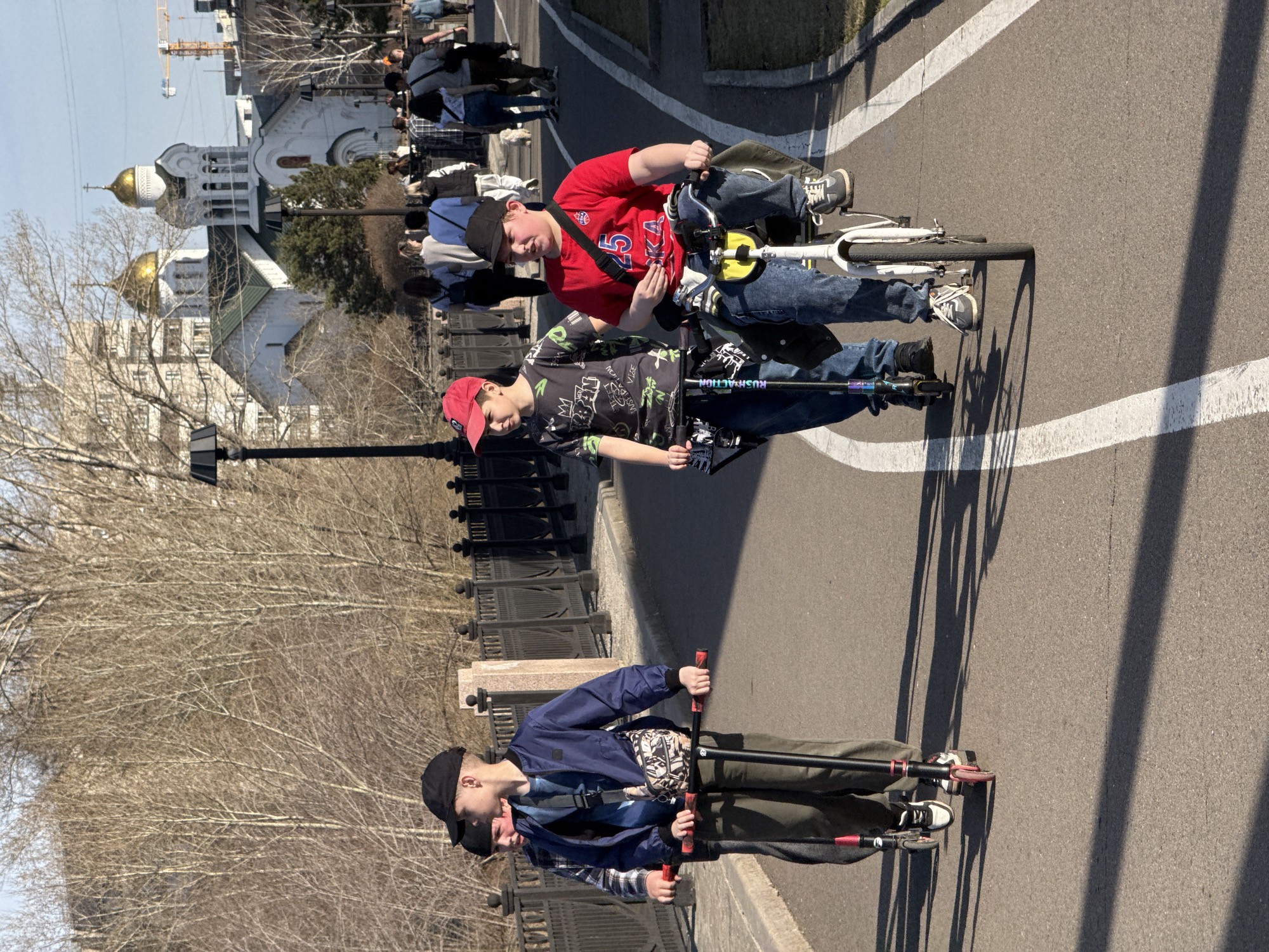Three young males are present on a paved path. One male is riding a white bicycle. Another is on a scooter. The third male is also on a scooter. They are close together on the path. The environment includes bare trees on one side and a metal fence. Beyond the fence is a building with golden domes, indicating an Orthodox church. People walk in the background, suggesting a public space. A street lamp is also visible near the fence. A construction crane is visible in the background. The photo appears to be taken during the day under sunny conditions. Based on the architecture of the church and the environment, the location is likely Krasnoyarsk, Russia.