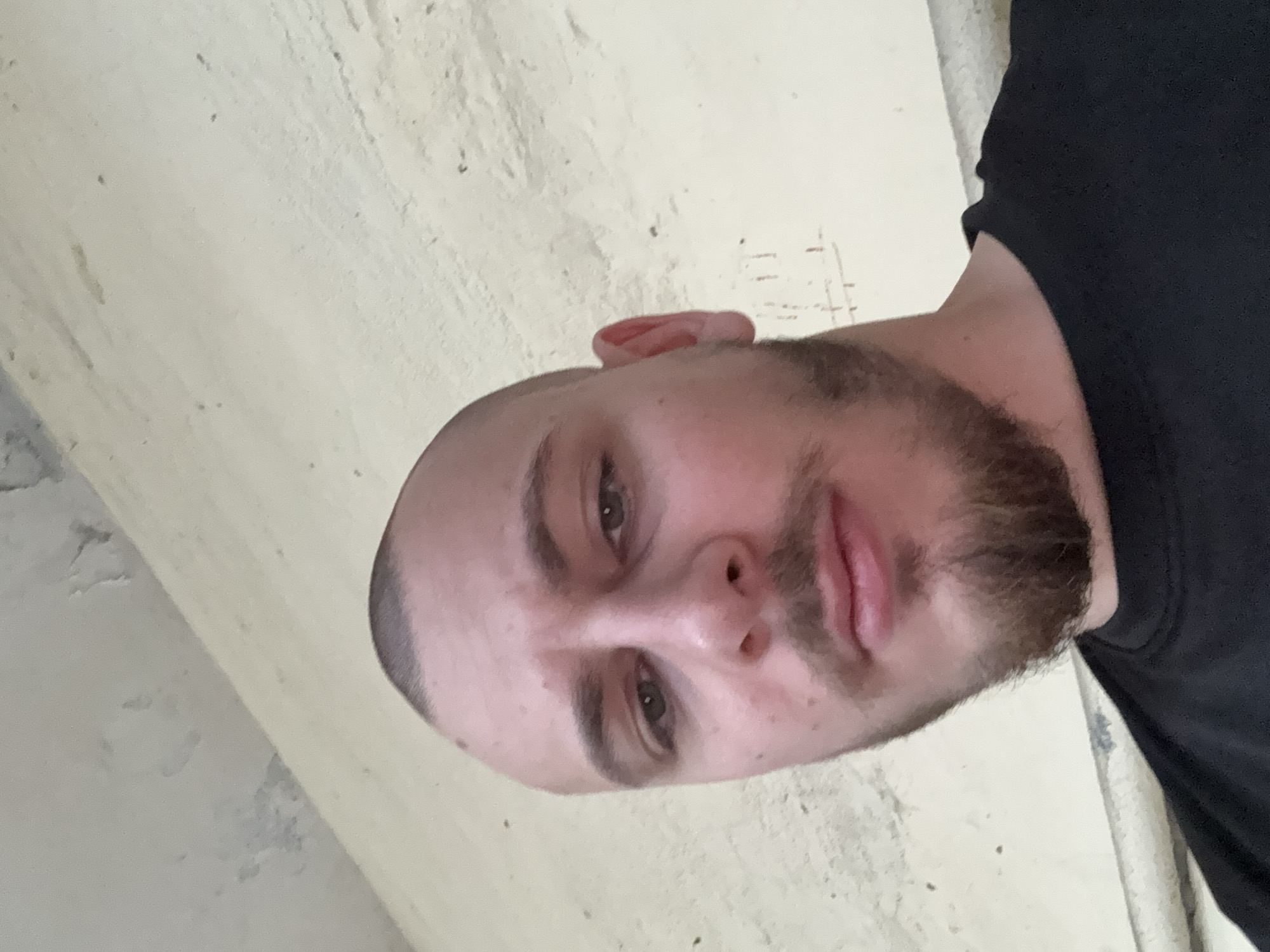 A close-up, tilted photograph shows a Caucasian man with a shaved head and a beard. He is wearing a black t-shirt. The background appears to be a plain, light-colored wall, and a corner of a wall or structural element can be seen. Without further context or location information, it is impossible to determine if the photograph was taken in Stara Zagora, Bulgaria.