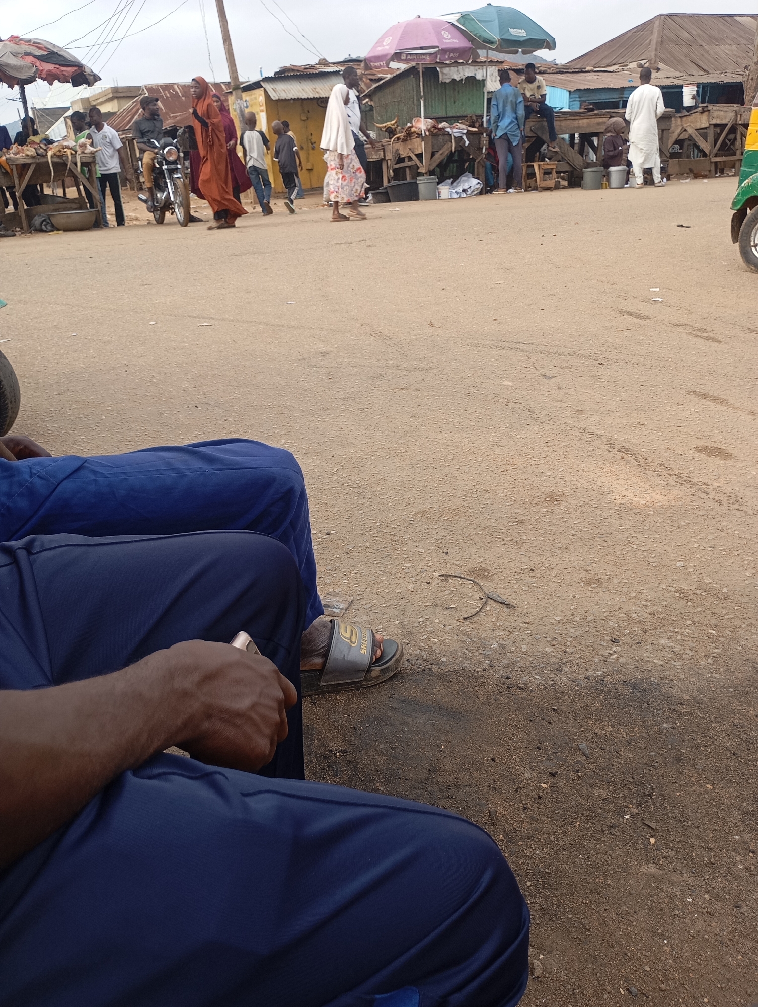 The image depicts a busy street scene in Jos, Nigeria, likely a marketplace given the presence of vendors and goods. In the foreground, two individuals are seated, their legs in dark blue trousers visible. One person's hand, holding a mobile phone, rests on their leg. The ground is a rough, sandy surface.
In the background, the marketplace is active with people. A man rides a motorcycle, and a woman in an orange headscarf and dress walks beside it. Several other individuals, including children, are scattered throughout the scene, some appearing to be shoppers or vendors. Stalls with umbrellas and awnings display goods, possibly food items like meat, as suggested by the visible carcasses on a table. Structures with corrugated metal roofs line the background, indicating a built-up urban environment. A three-wheeled vehicle, commonly known as a "Keke Napep" in Nigeria, is partially visible on the right side of the image, suggesting transportation is readily available. The overall atmosphere is one of daily commerce and activity in a Nigerian city.