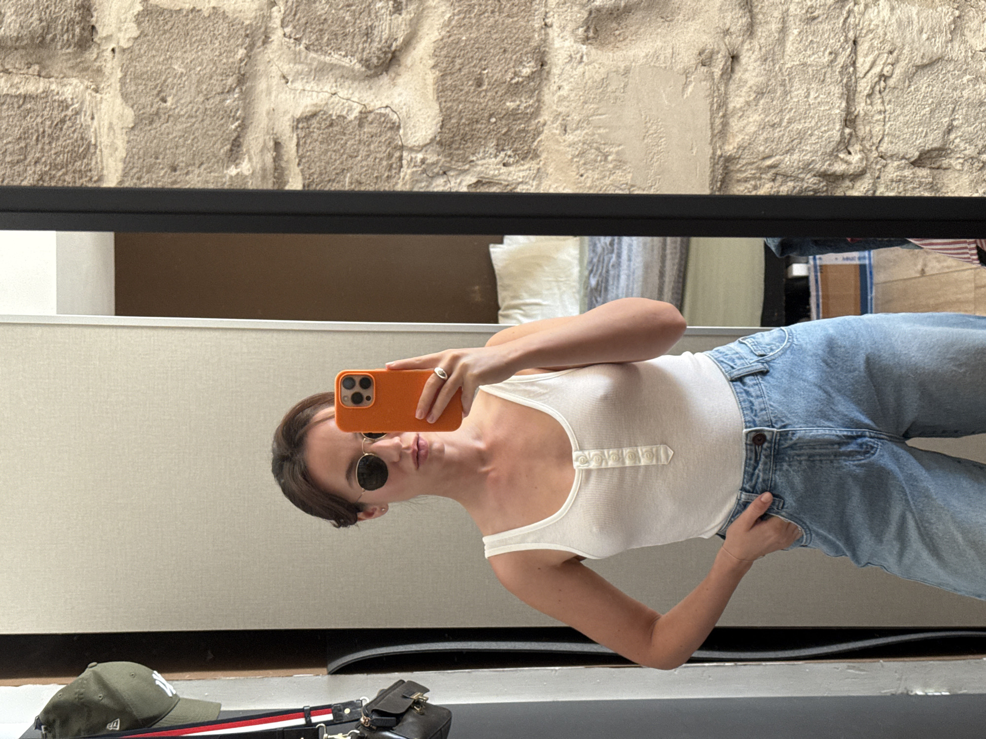 A woman is taking a mirror selfie in a room with a stone wall. She is wearing a white ribbed tank top with buttons down the front and light blue wide-leg jeans. She has on round sunglasses and a silver ring on her right hand. Her orange-colored phone is visible as she takes the picture. In the reflection, part of a bed and a closet door are visible. A green baseball cap hangs on a hook on the closet door. The scene appears to be indoors in an apartment.