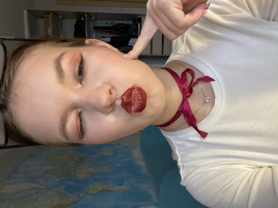 A young female subject is depicted in a close-up, indoor setting within the city of Chaikovsky, Russia. She has light skin, light brown hair pulled back, and light brown eyebrows. Her left eye is winking, and her lips are puckered into a "kiss" formation, accentuated by glossy, dark red lipstick. Pinkish-red eyeshadow is visible on her eyelids. Her right index finger is pressed against her right cheek. She wears a white, crew-neck t-shirt. A dark pink/fuchsia satin ribbon is tied in a bow around her neck, functioning as a choker. Below this, a thin silver chain necklace with a small, square-cut, clear gem pendant is visible. The frames of eyeglasses are positioned on top of her head.
The background on the left features a portion of a wall-mounted world map, predominantly blue for oceans and various lighter shades for landmasses. Below the map, a light blue object, possibly a piece of furniture, is partially visible. On the right, an open closet or shelving unit made of light-colored wood or laminate displays folded clothing and a few boxes on its shelves. The lighting in the scene is soft and ambient.