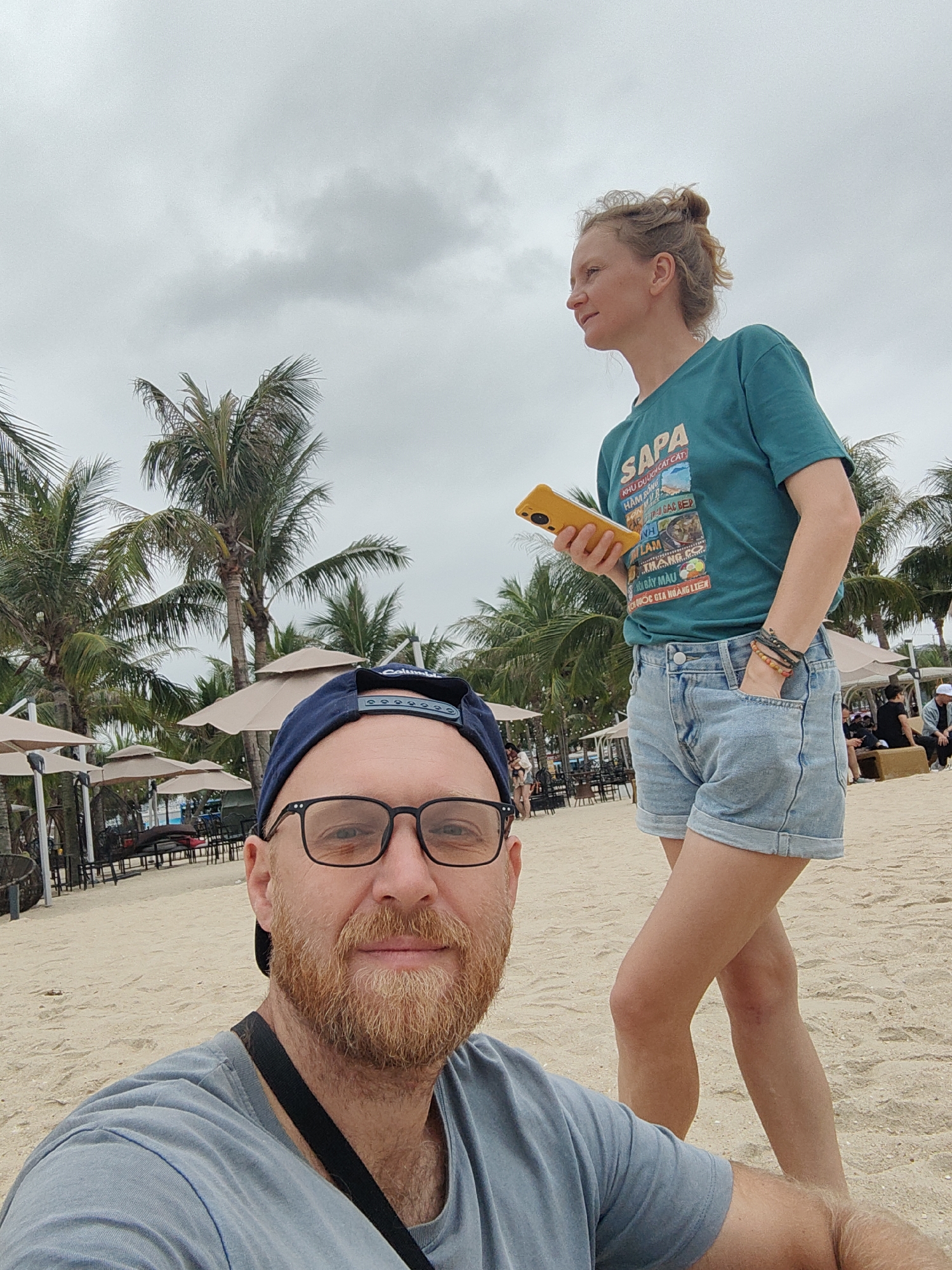 The image captures a selfie of a fair-skinned man with a reddish beard, wearing a blue baseball cap and glasses, against a backdrop of a beach scene. Behind him stands a fair-skinned woman with her hair up, holding a yellow phone and wearing a turquoise shirt with the word "SAPA" on it and denim shorts. The location appears to be a beach, with sand visible in the foreground and rows of palm trees and umbrellas in the background. The image was taken in Bai Chay Ward, Vietnam.