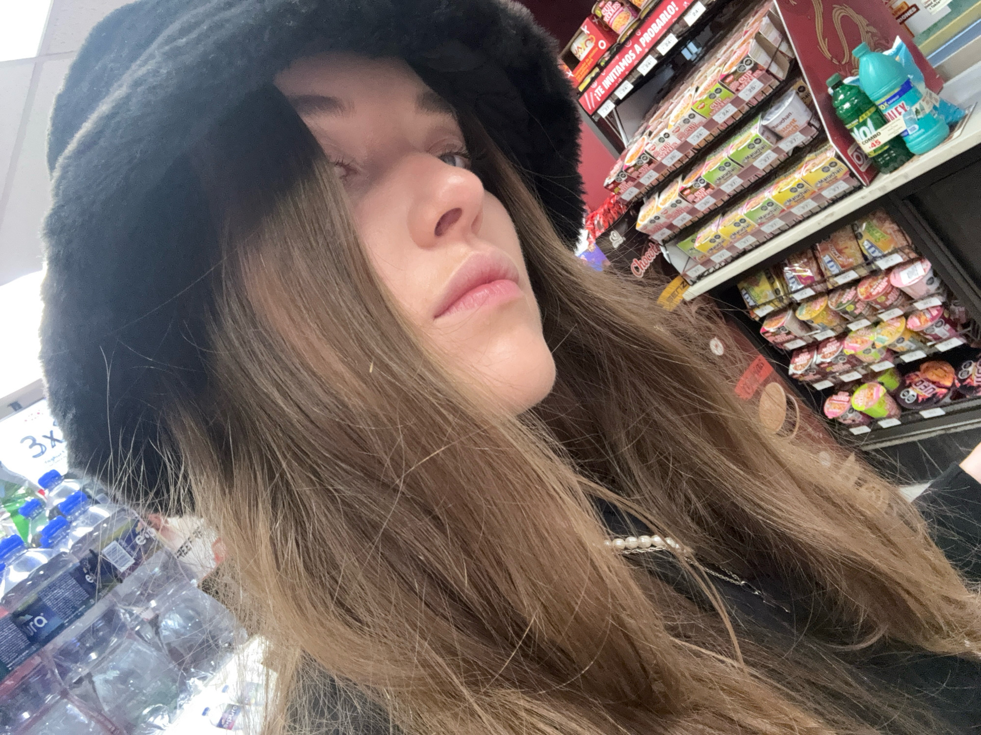 A woman wearing a black fuzzy hat poses for a selfie in what appears to be a convenience store or small market in Mexico City, Mexico. She has long, light brown hair and is wearing a pearl necklace. Behind her, shelves are stocked with various products, including bottled water, instant noodles, and cleaning supplies. The scene is brightly lit, and the focus is on the woman's face and upper body.