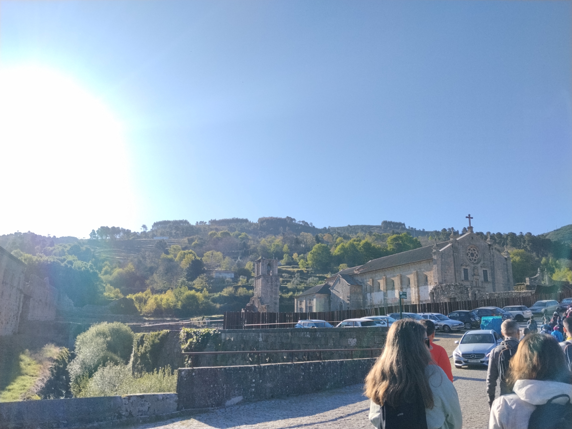 The image depicts the Monastery of Saint John of Tarouca in Tarouca, Portugal. The stone monastery, with a cross atop, stands behind a stone bridge and a parking area filled with various cars. A bell tower is partially visible to the left of the monastery. In the foreground, people are walking on the stone bridge, with their backs to the viewer. The landscape features trees and terraced hills behind the monastery. The sky is clear and blue, with the sun shining brightly.