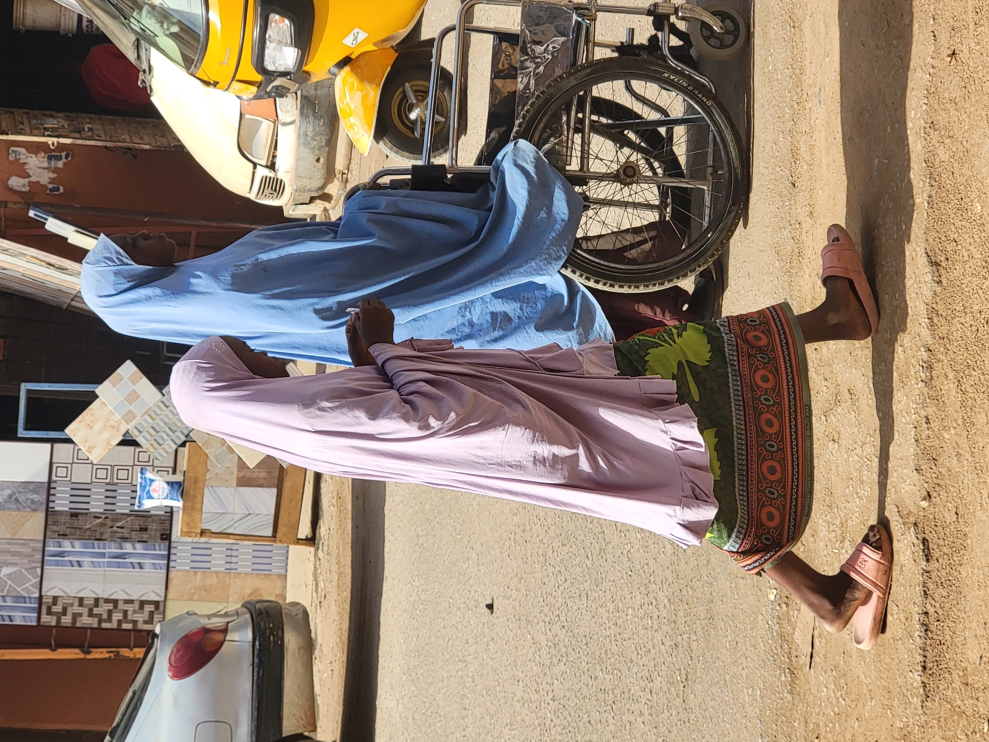 The image captures a sunlit street scene in what appears to be a commercial area, consistent with Maiburiji, Nigeria. Two individuals, likely women, are the central subjects. One is dressed in a light blue, flowing garment and head covering, positioned next to or in a wheelchair. Her back is partially to the camera, and only a portion of her face is visible, looking towards the left. The other individual, closer to the foreground, wears a lavender-pink long-sleeved top and head covering, paired with a vibrant green, orange, and red patterned skirt or wrapper. She is standing or sitting, with her right foot, clad in a pink slip-on sandal, visible on the sandy ground.
In the background, a yellow auto-rickshaw or "keke Napep" is parked or driving, with its front and side visible. A silver car's rear bumper and taillight are visible on the far left. A wheelchair, bearing the text "KYLIN BRAND" on its tire, is prominent between the two individuals. To the left of the person in lavender, a display of various patterned ceramic tiles is set up, possibly indicating a tile shop. A small weighing scale is perched on this display.
The bright sunlight casts long shadows on the ground, suggesting it is daytime, likely morning or late afternoon. The overall impression is a candid snapshot of daily life in an urban or semi-urban environment. There is no specific event or interaction discernible, merely people present in their surroundings.