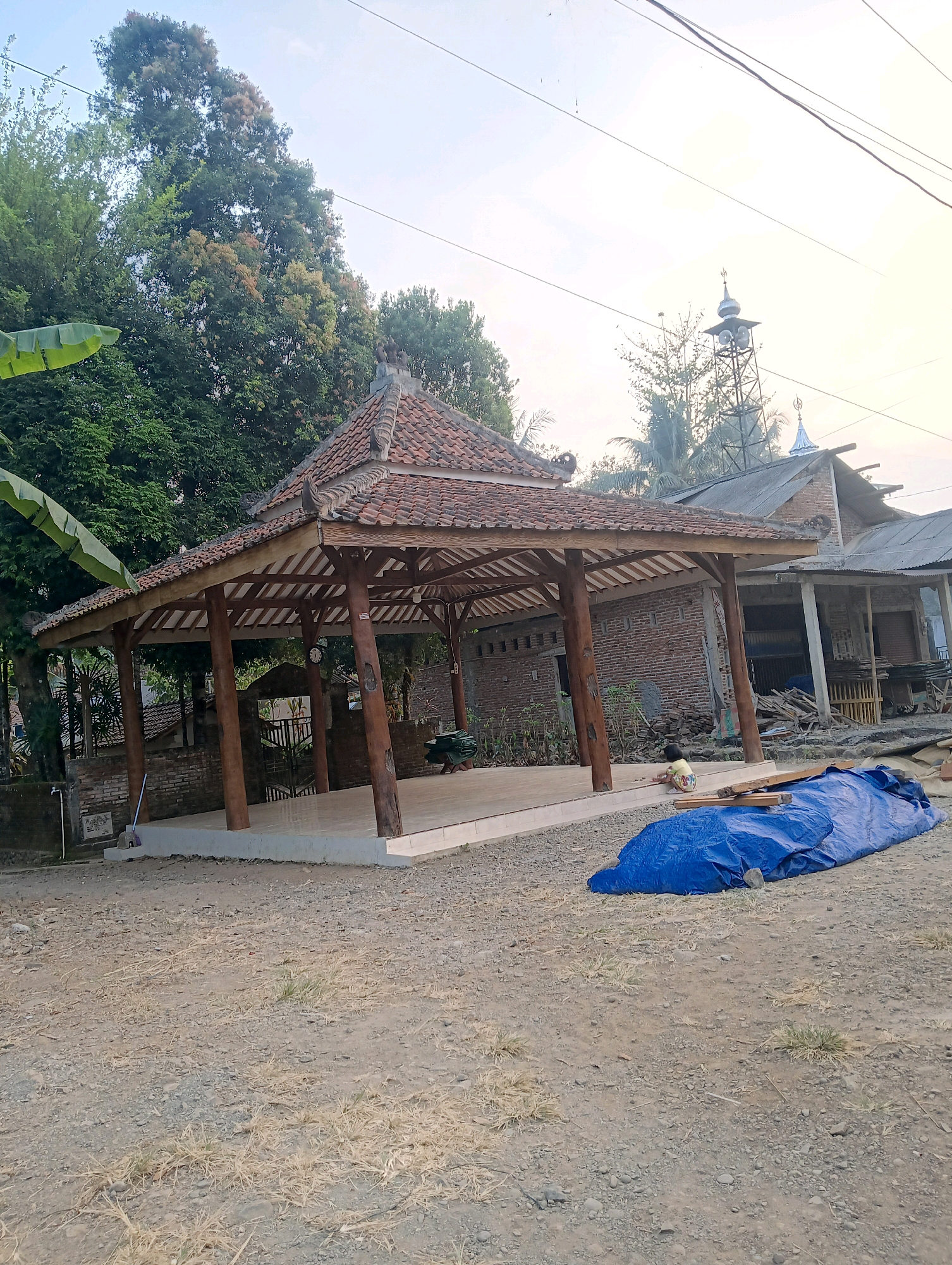 In the village of Kedawung, Indonesia, a wooden gazebo with a tiled roof stands prominently in a dirt lot. The gazebo features thick wooden posts supporting the roof and an open, tiled floor. To the right of the gazebo, a blue tarp is stretched over some unseen objects, with a child in a yellow shirt and patterned shorts sitting near it, engaged in an activity. Behind the gazebo, a brick building is visible, with a portion of its roof extending forward. Further in the background, a tall metal structure, possibly a minaret or a loudspeaker tower for a mosque, pierces the sky, with a glimpse of a mosque dome. Lush green trees surround the area, suggesting a rural or semi-rural setting. The ground is uneven and covered with dirt, gravel, and sparse dry grass, indicating an outdoor environment under a bright, overcast sky.