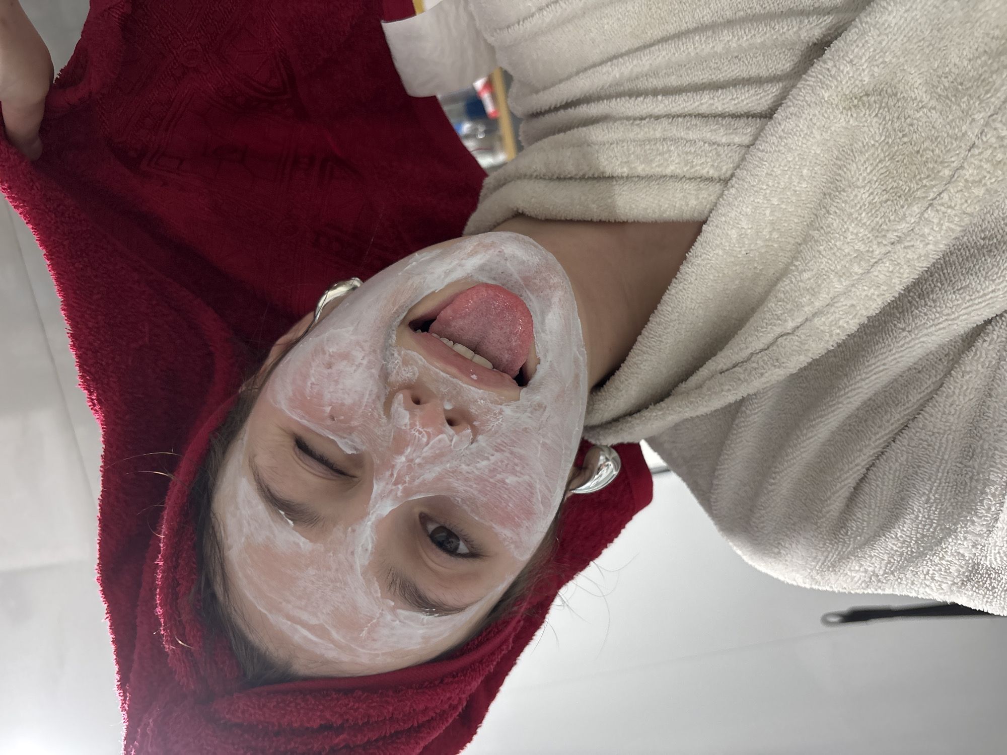A person is lying down, facing upwards, with a white facial mask on and their tongue sticking out. They are wearing a red towel on their head and a white terry cloth robe. The location is Фоки, Russia.