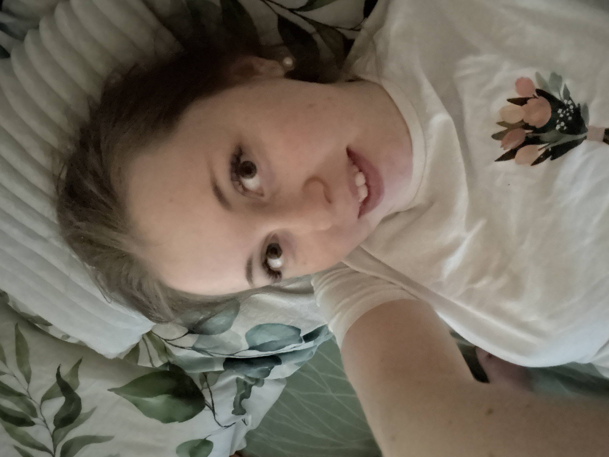 A young woman is lying on her back on a bed. The bed has a white, possibly floral-patterned, duvet. She is wearing a white shirt with a floral design on the upper right side. Her hair is brown and pulled back. She is looking directly at the camera with a slight smile. The location is unknown.