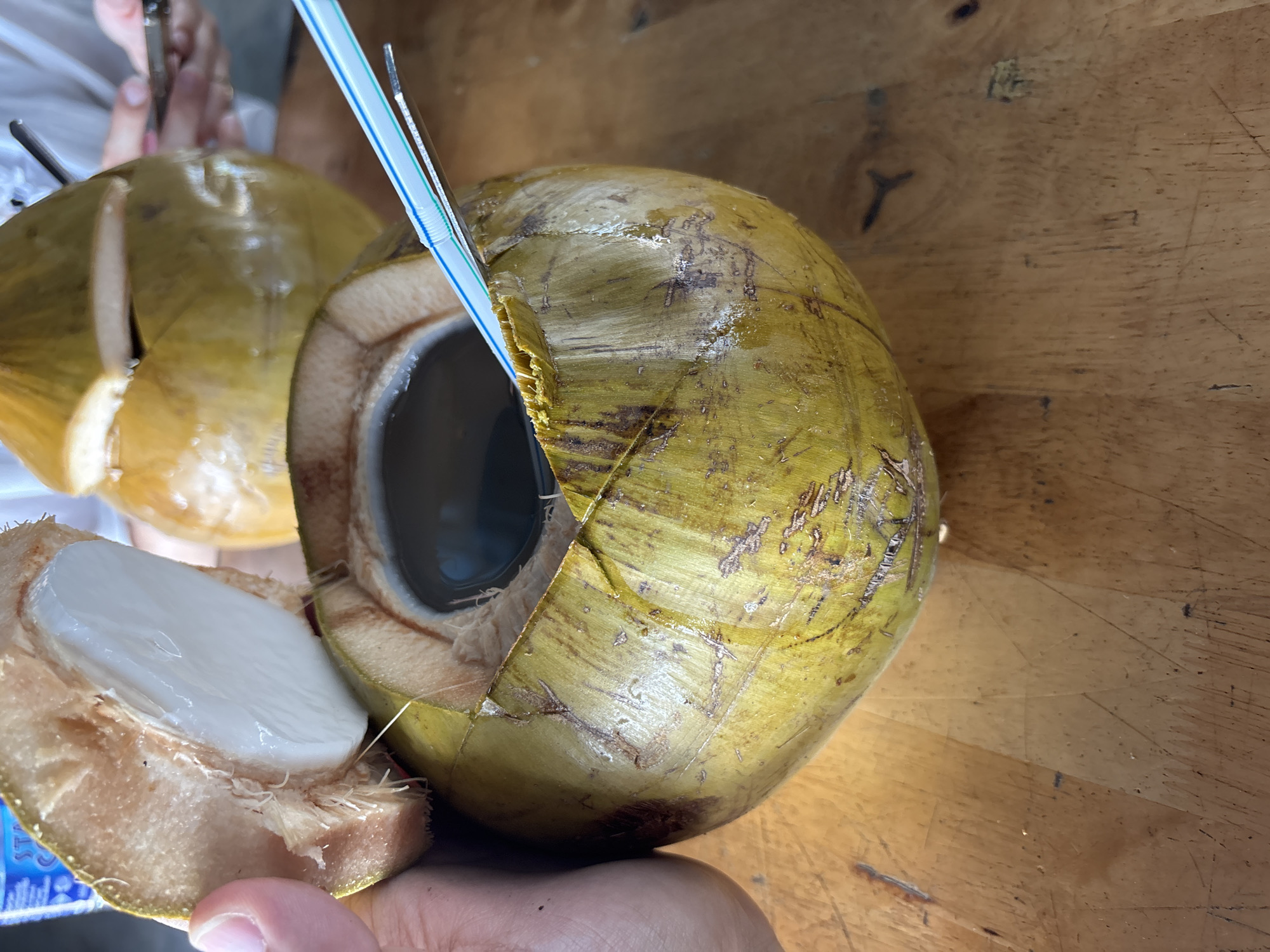 In George Town, Malaysia, this image captures a close-up view of a young, green coconut being held open, revealing its white flesh and clear coconut water. A blue and white striped straw is inserted into the coconut water. In the background, another whole green coconut is visible, with a metal spoon resting on its opened top. The scene takes place on a wooden table. A person's hand is on the left, holding the opened coconut. In the blurred background, another individual is partially visible, appearing to be holding a mobile phone. The lighting suggests an indoor setting, possibly a cafe or restaurant.