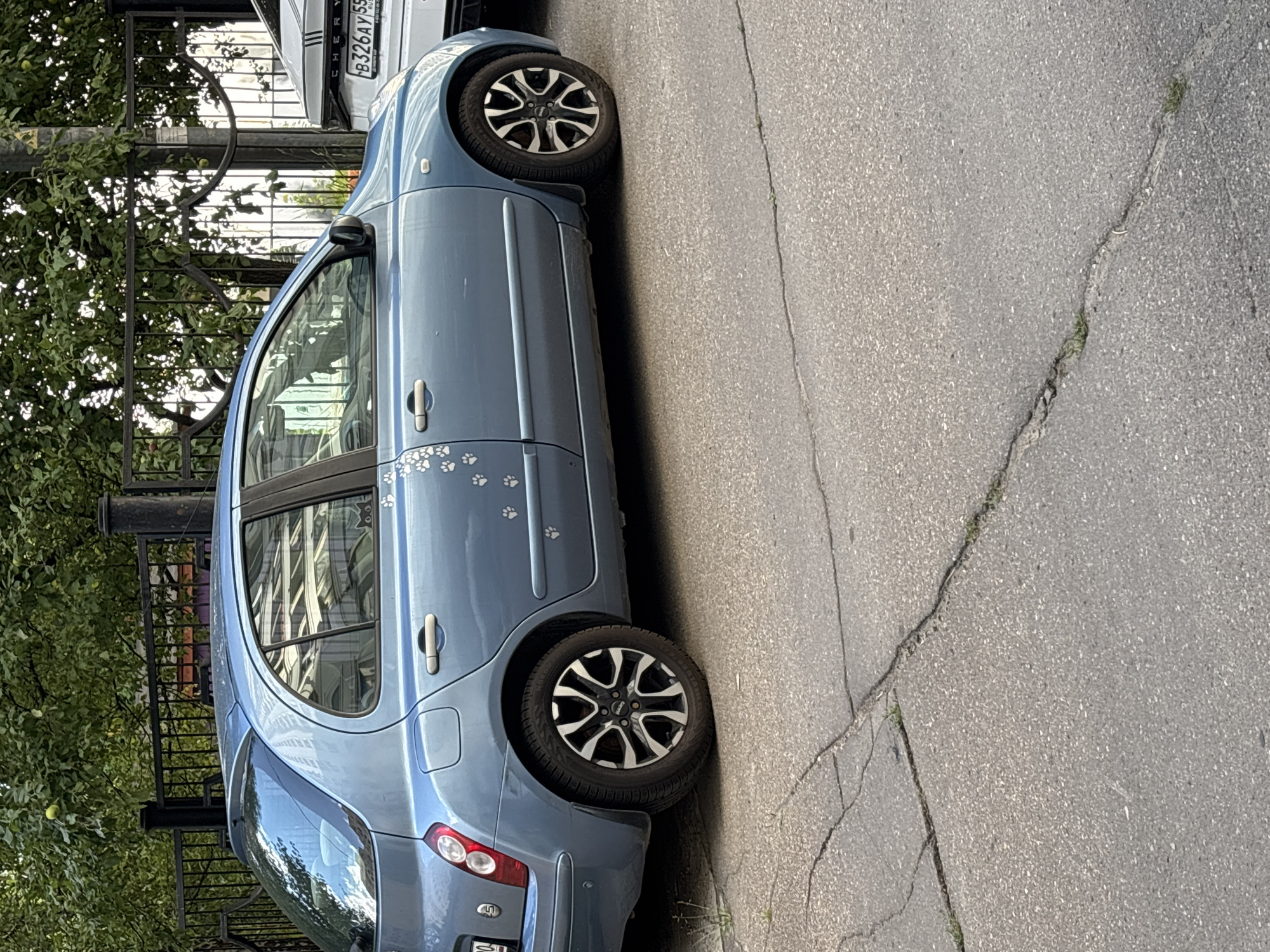 A light blue Nissan hatchback is parked on a cracked asphalt surface in Odintsovo, Russia. The car has several white paw print decals on its side, with a small black cat silhouette sticker on the rear passenger window. The car is partially obscured by a black metal fence and trees with green foliage in the background. To the right of the Nissan, a portion of a white SUV with a Russian license plate is visible. The scene appears to be a residential street or parking area.