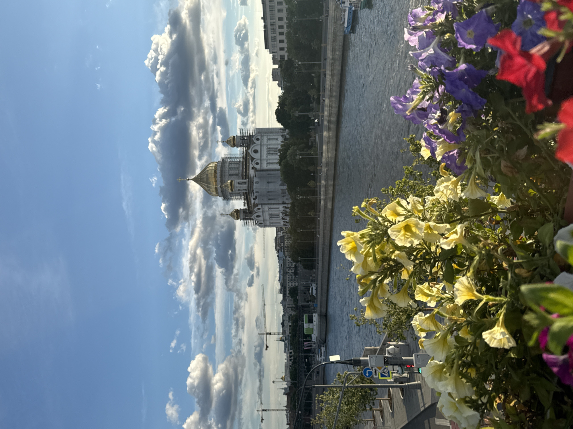 This media file captures a vertical view of the Moskva River and the Cathedral of Christ the Saviour in Moscow, Russia. In the foreground, a profusion of bright yellow and purple petunias spills over the edge of a planter, partially obscuring the view. The river flows calmly, reflecting the sky.
Across the river, the white walls and golden domes of the Cathedral of Christ the Saviour stand out, with scaffolding visible on one side, suggesting ongoing construction or renovation. Beyond the cathedral, a cityscape of buildings and trees lines the embankment. Several cranes are visible in the distance, hinting at ongoing development. A boat travels along the river, and on the street to the right, traffic lights and street signs are visible.
The sky is a vibrant blue, punctuated by dramatic, white cumulus clouds. The lighting suggests it is daytime, likely late morning or afternoon, with the sun casting clear shadows. The overall atmosphere is serene, with the natural beauty of the flowers and the grandeur of the cathedral creating a picturesque scene. No discernible text is visible in the image.